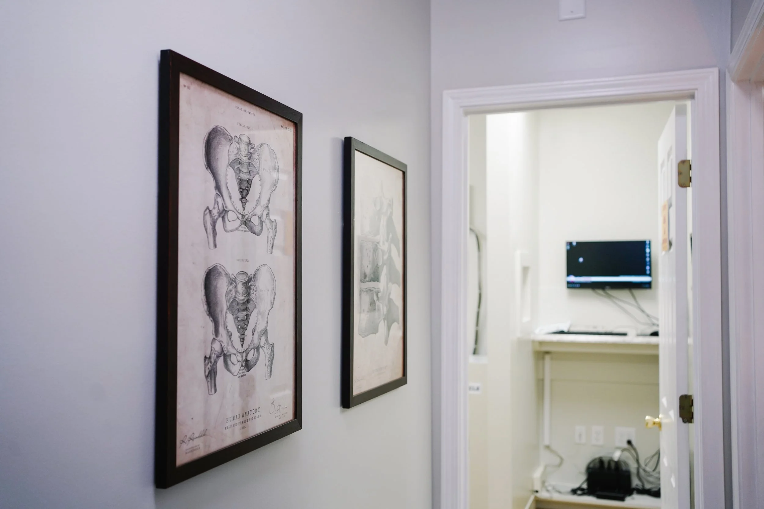 Hallway with framed anatomical hip diagrams and a room with a wall-mounted monitor and electronic equipment in the background.