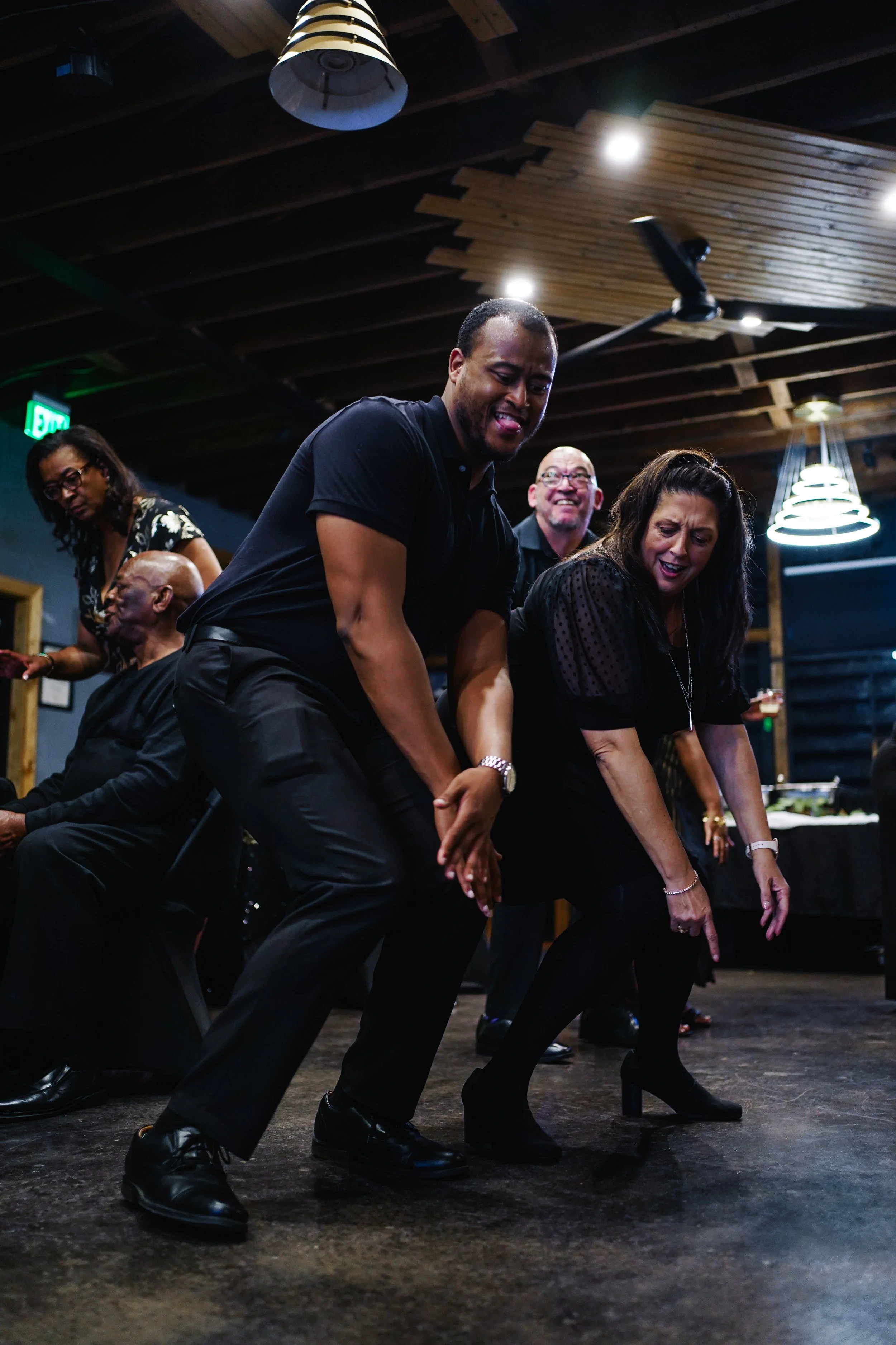 People dancing and enjoying themselves at a social gathering in a dimly-lit indoor venue with a wooden ceiling.