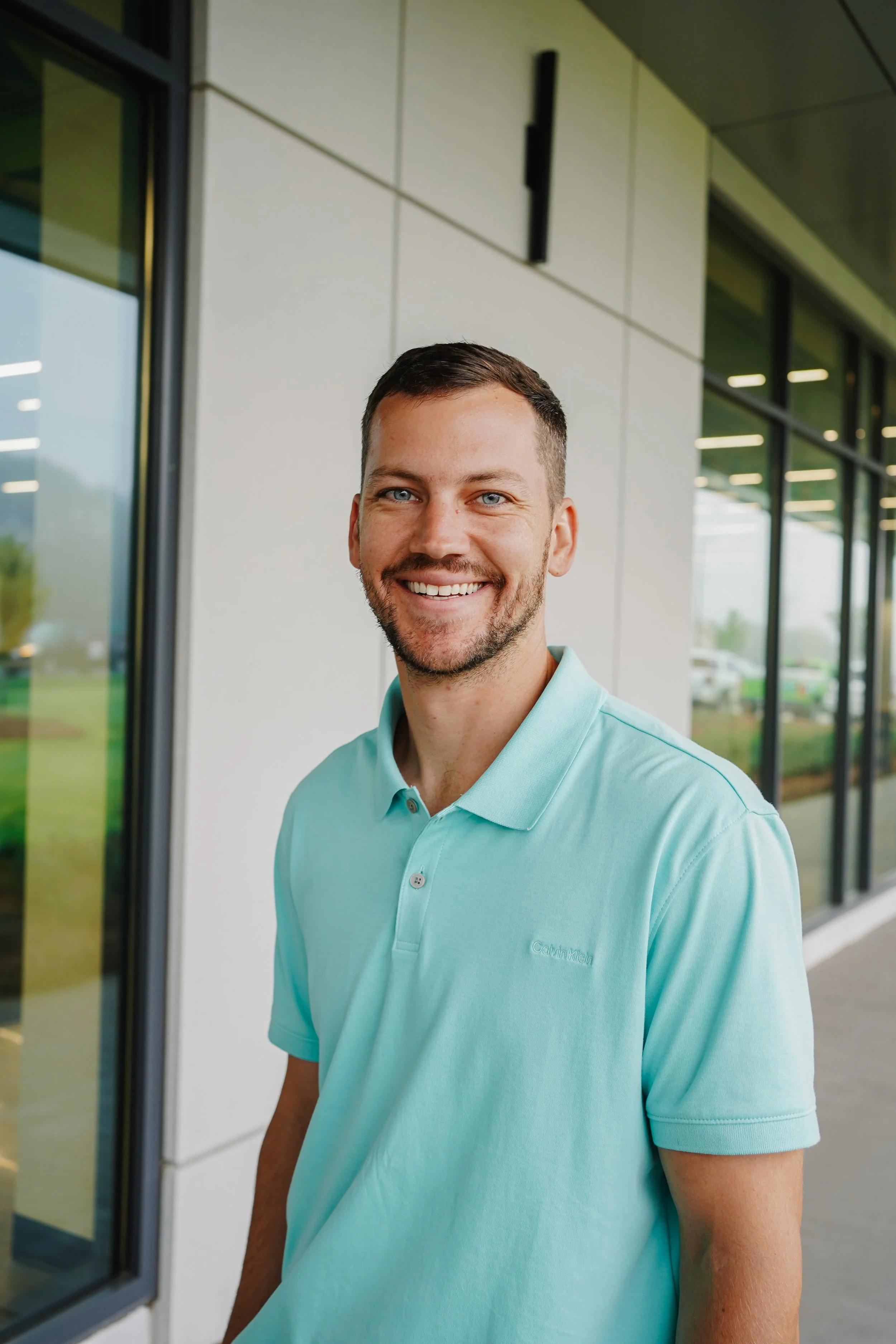 Smiling man in a mint green polo shirt standing outside of a modern building with large windows.