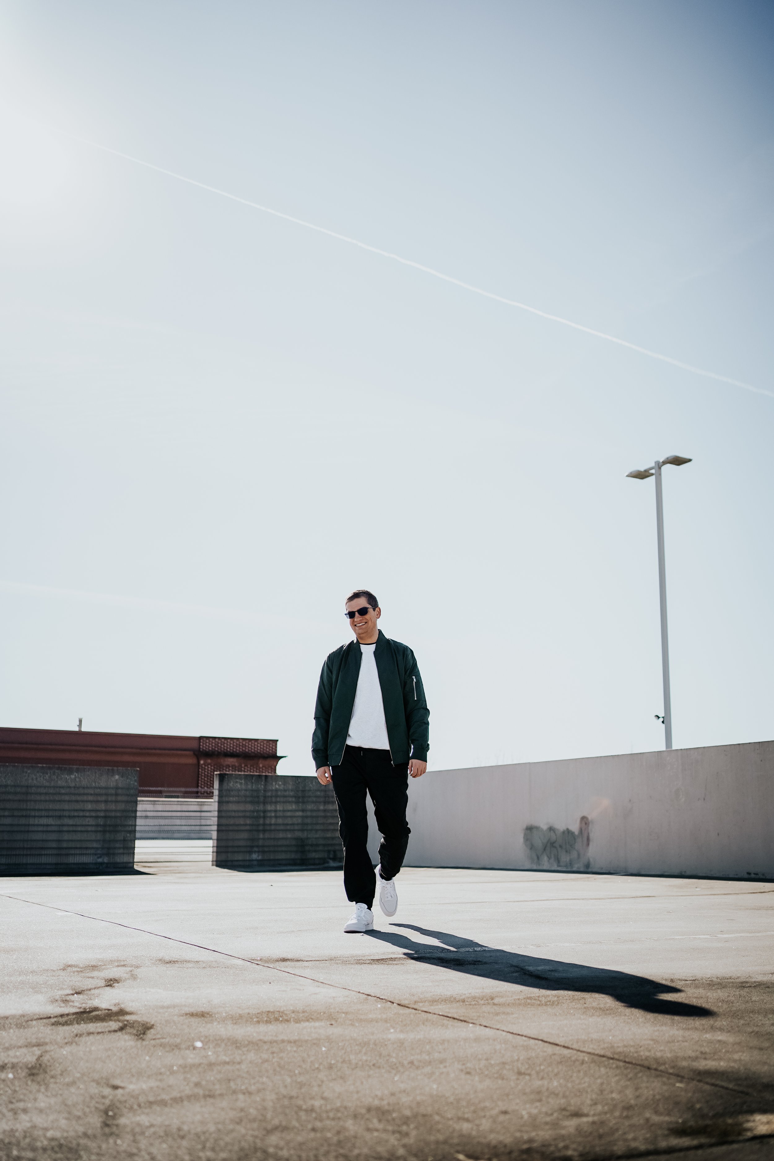 A man wearing sunglasses walking on a rooftop parking lot with minimal structures and a clear sky with contrails.