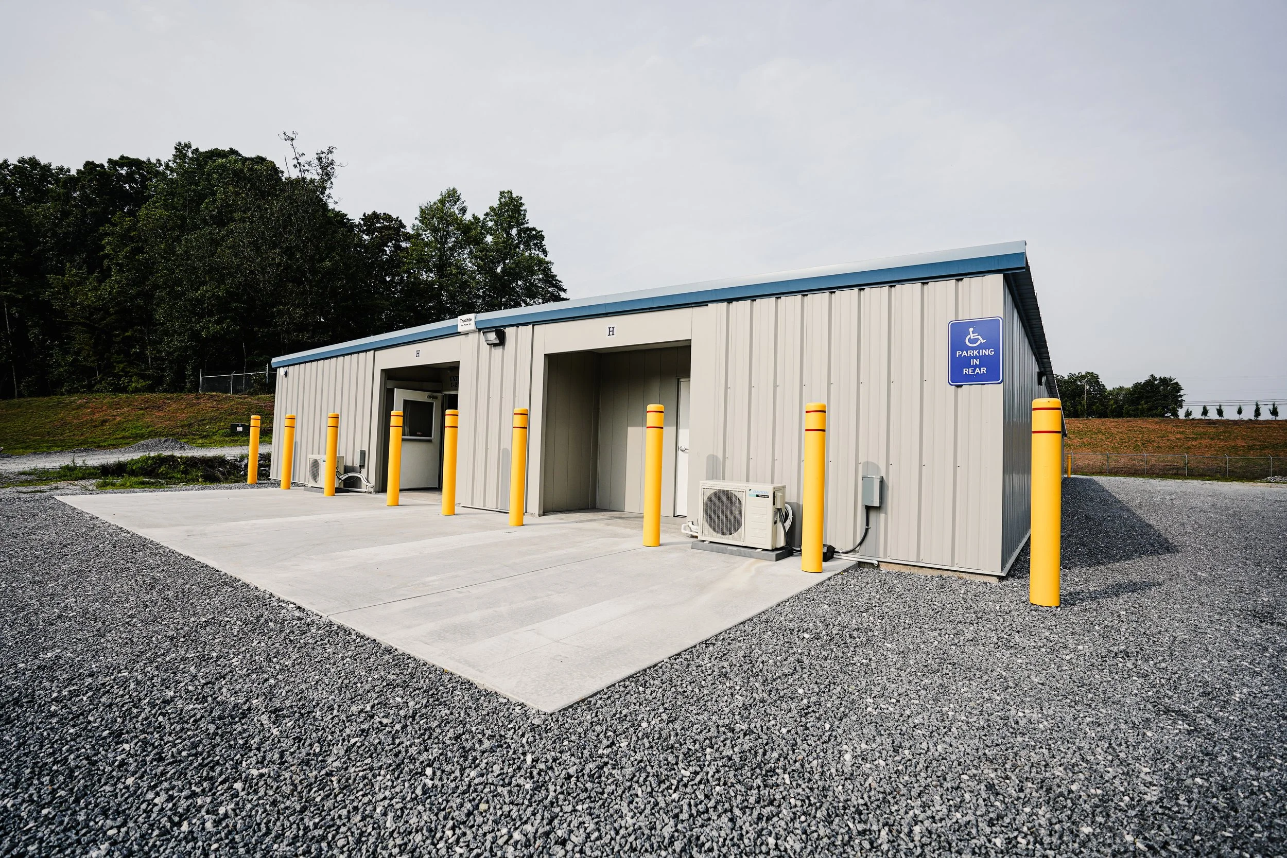 Small utility building with parking spots, including a designated handicap parking space, yellow post barriers, and an air conditioning unit outside, surrounded by gravel and trees in the background.