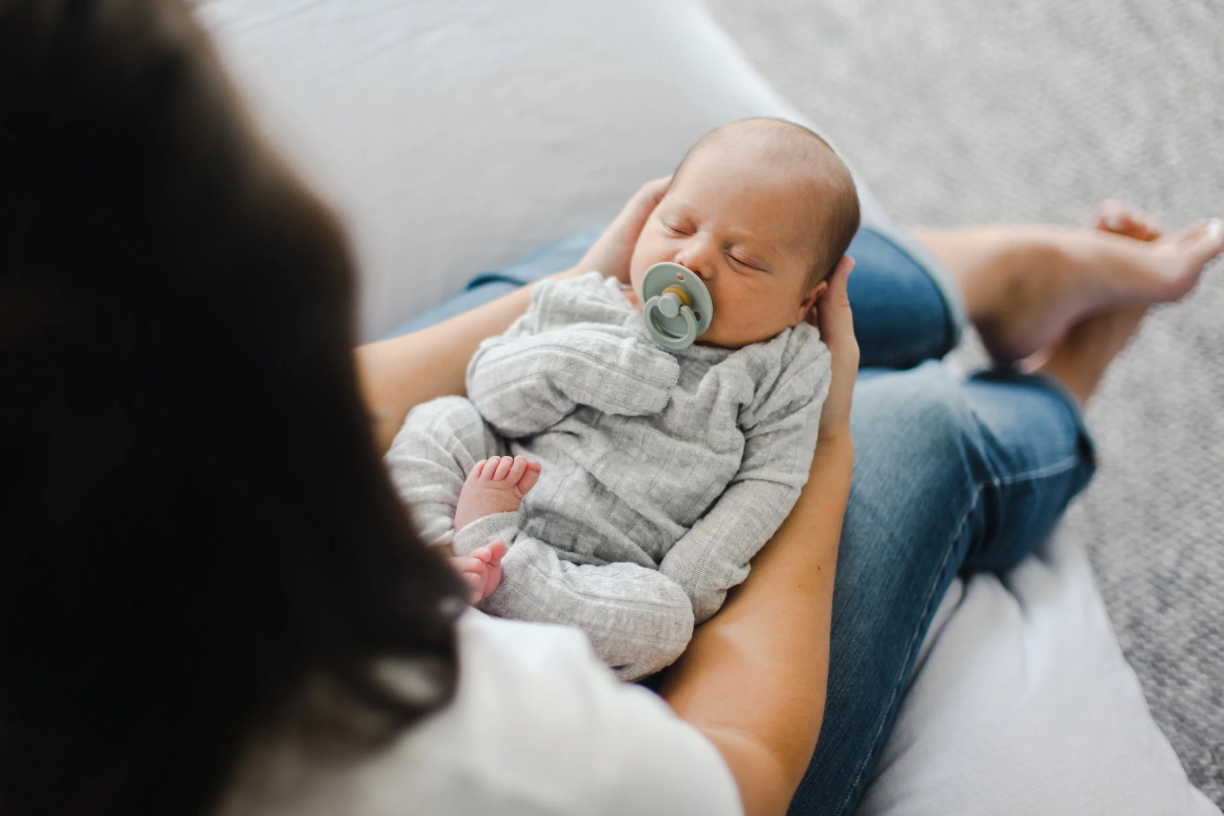 A person sitting on a sofa holding a sleeping baby with a pacifier, baby wearing a gray striped outfit and the person wearing blue jeans and a white top.
