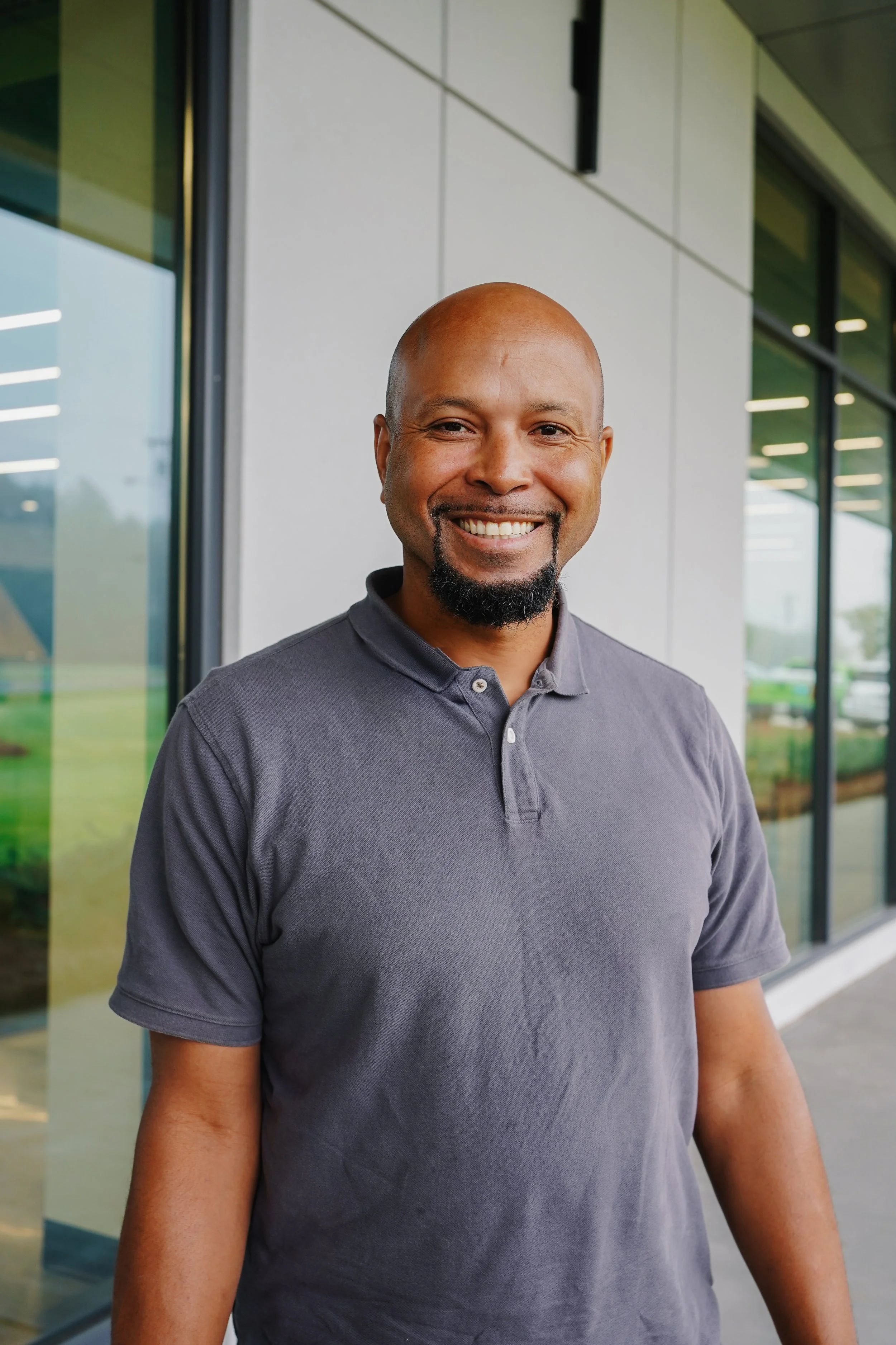 A smiling man with a beard and bald head standing outside a modern building with glass windows.