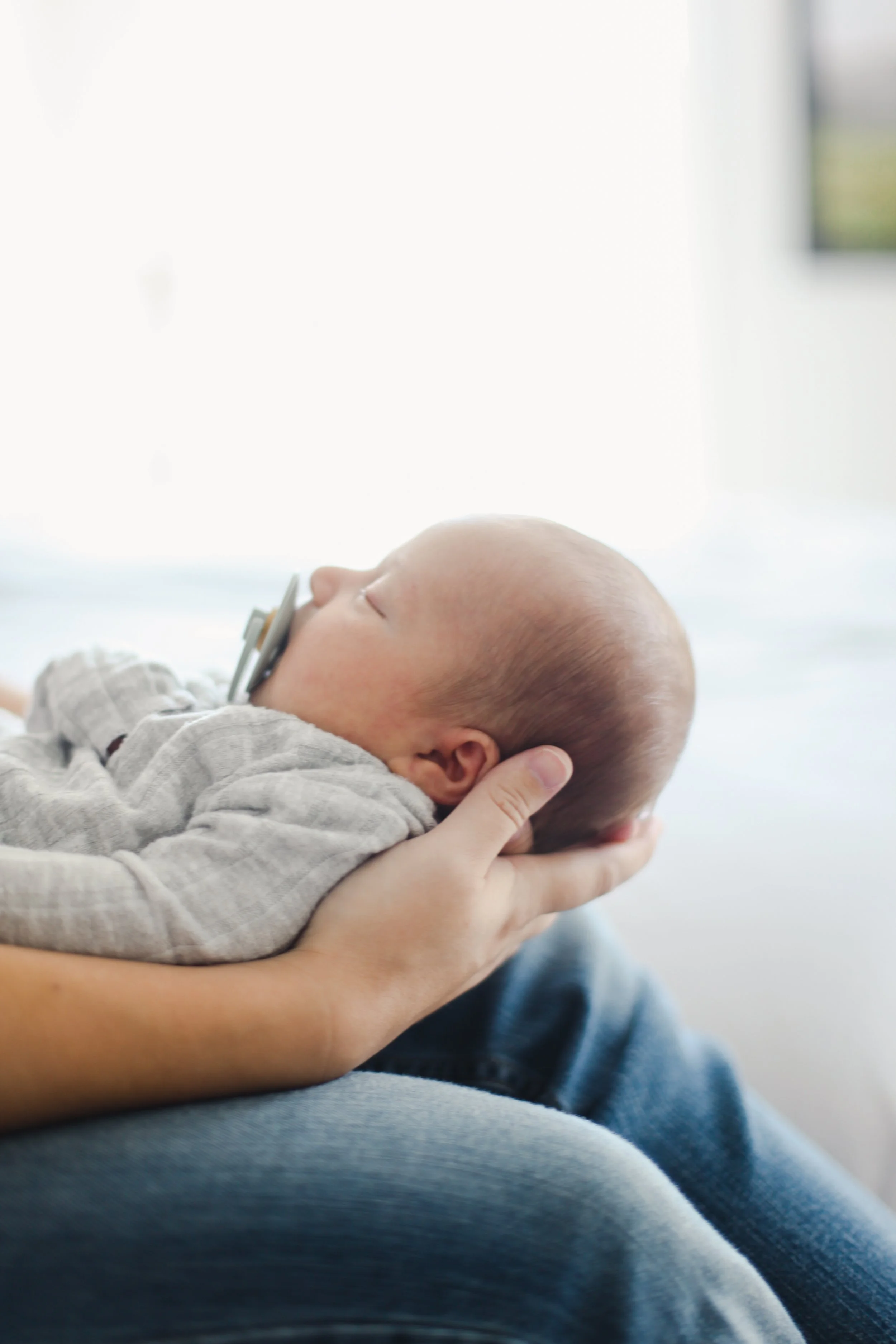 A person is gently holding a sleeping baby with a pacifier in its mouth, resting on their lap.