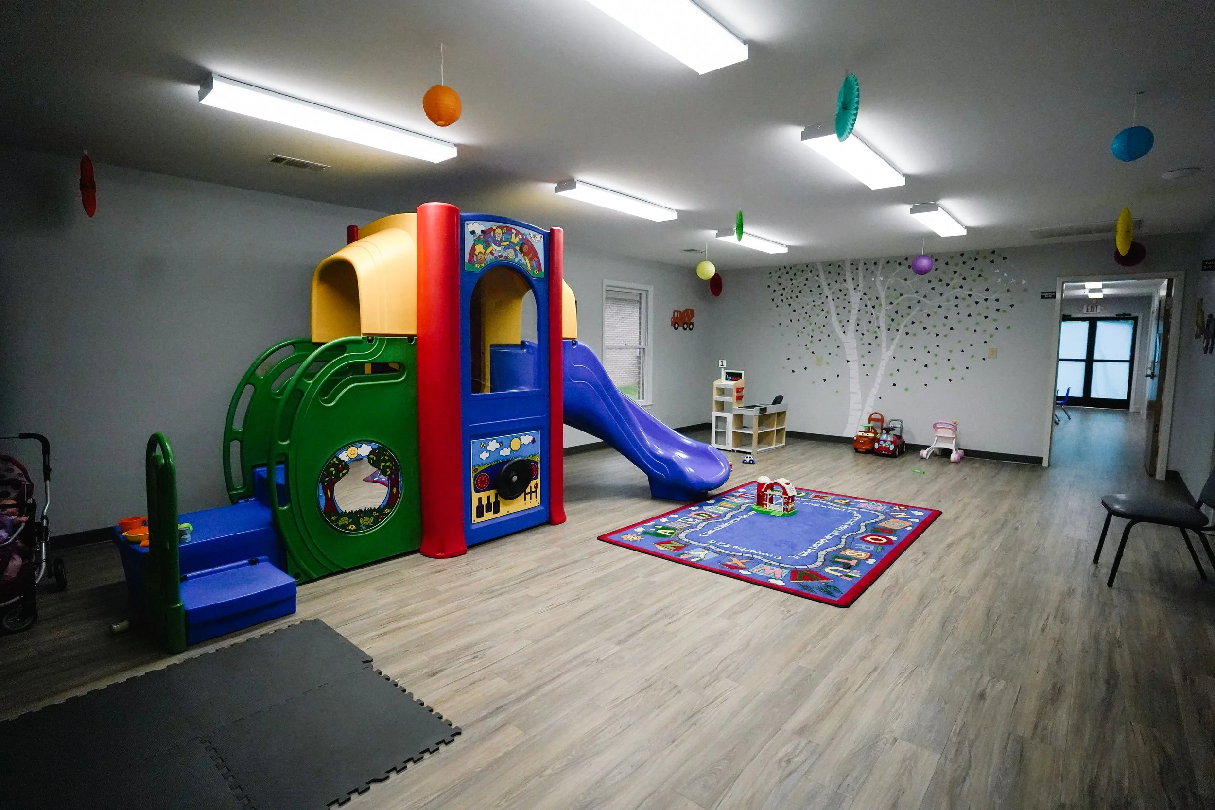 Indoor children's playroom with a colorful plastic play structure, slide, and various toys, decorated with hanging paper lanterns and a painted tree on the wall.