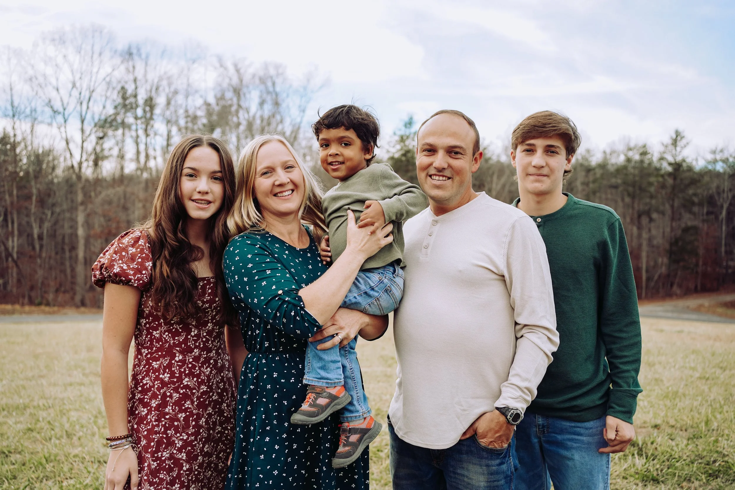 A family of five standing outdoors on a grassy field with trees in the background, smiling at the camera.