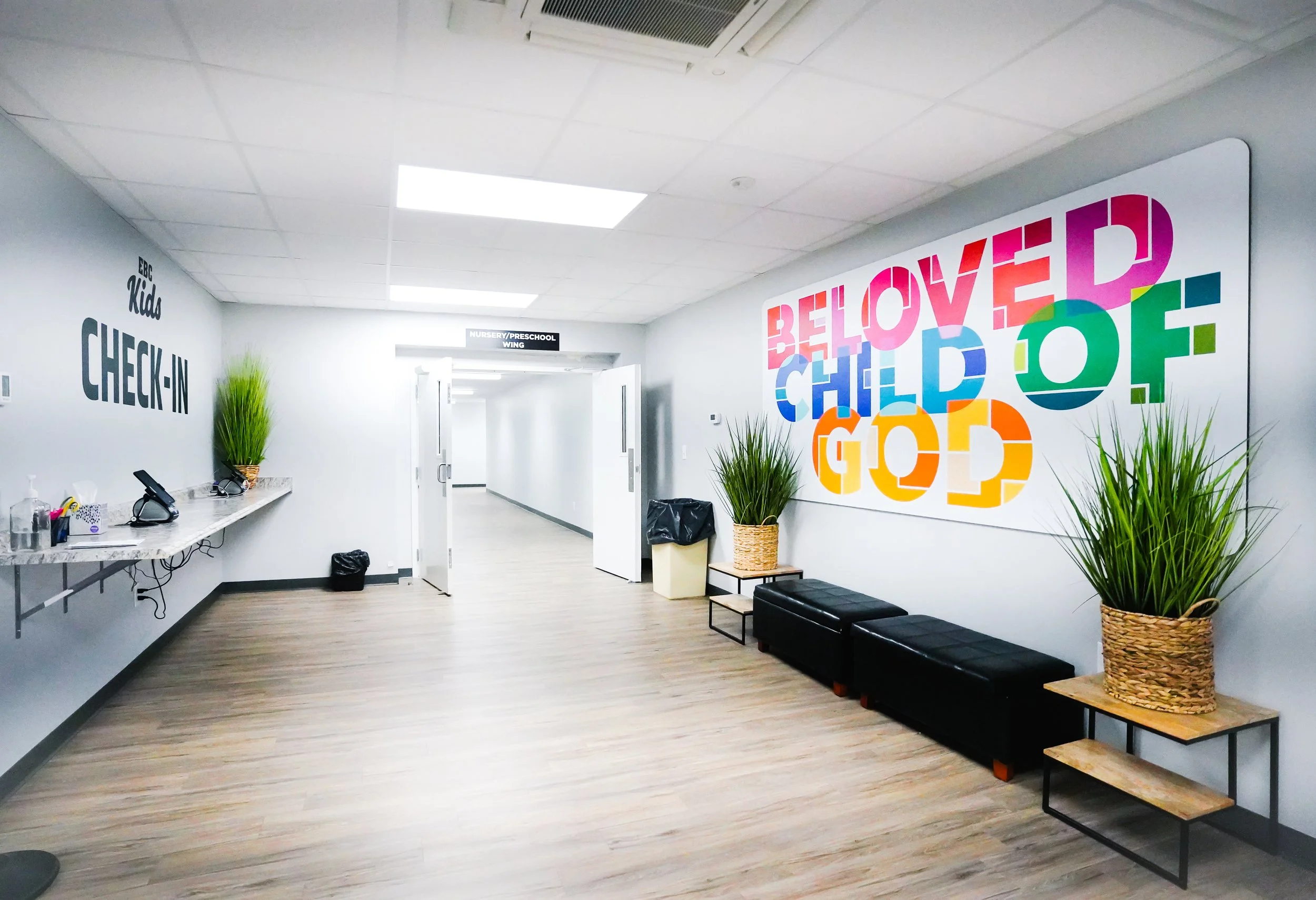 Lobby area with check-in counter, potted plants, benches, and colorful wall art with the words 'Be loved child of God'.