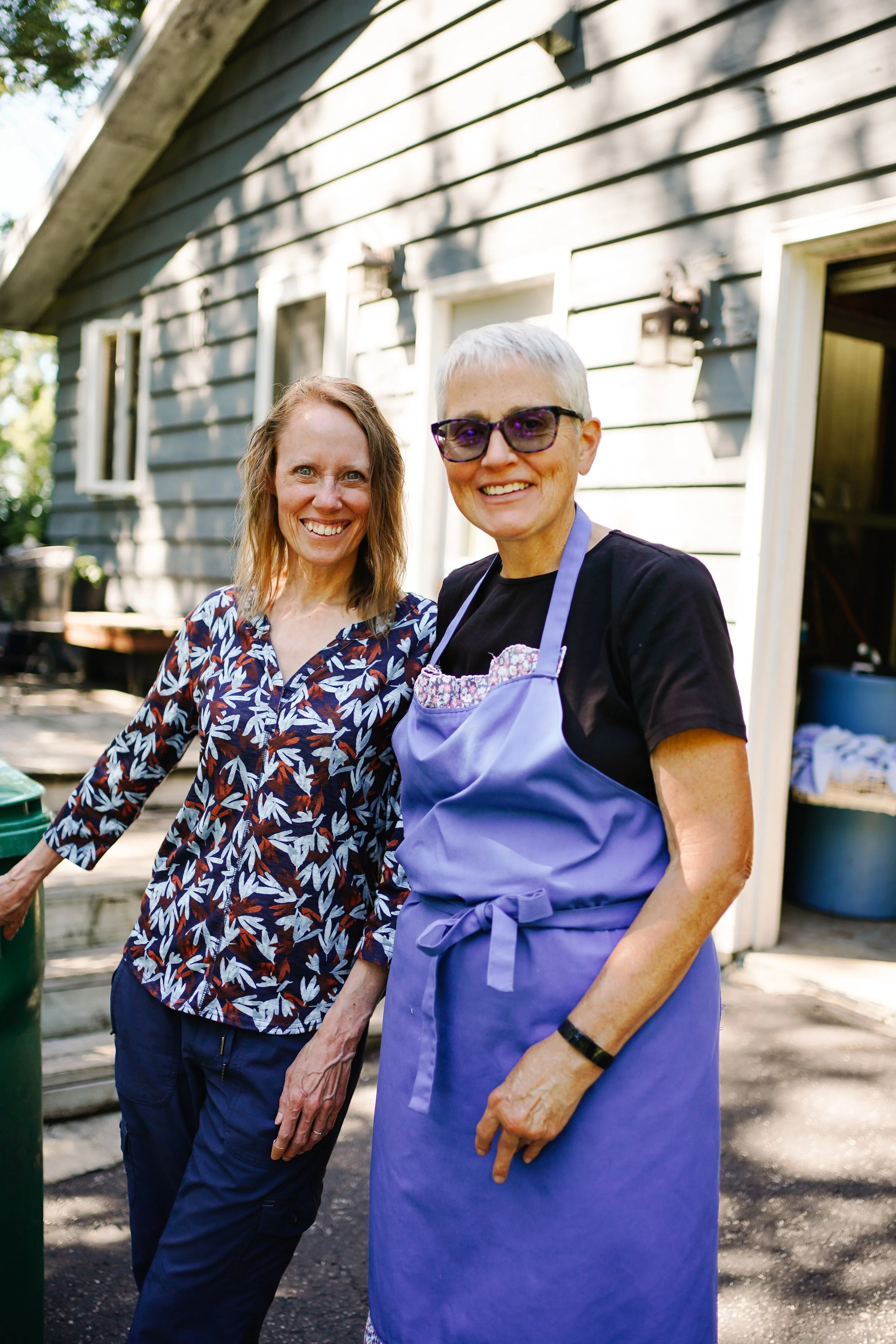 Two women standing outdoors in front of a gray house, smiling at the camera. One woman is wearing a floral shirt and the other is wearing a black shirt, purple apron, and glasses.
