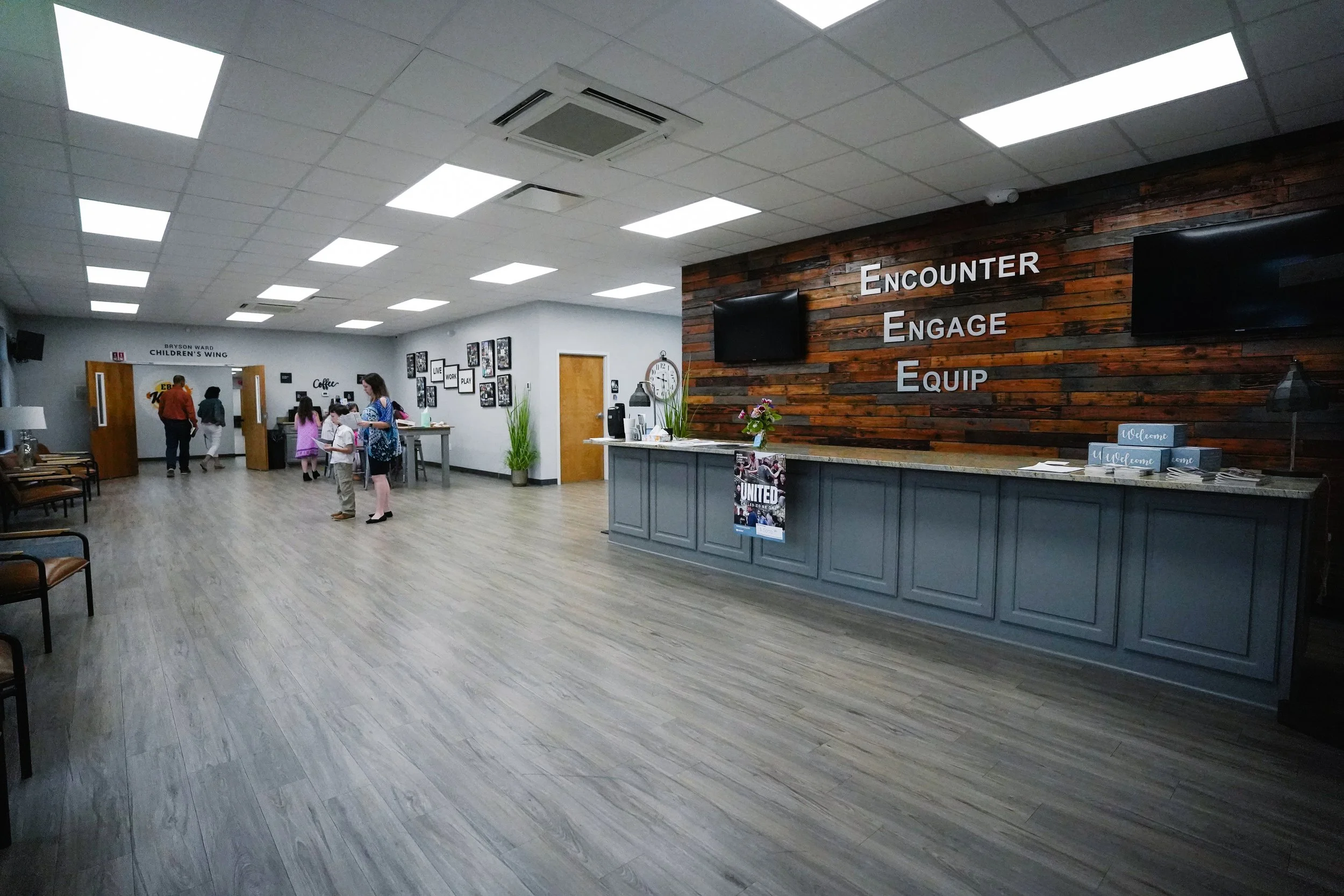 Lobby area with a wooden wall behind the reception desk displaying the words 'Encounter, Engage, Equip'. Several people, including children, are in the background near the doorway labeled 'Bryson Ward Children's Wing.' The floor is wood, and the ceil