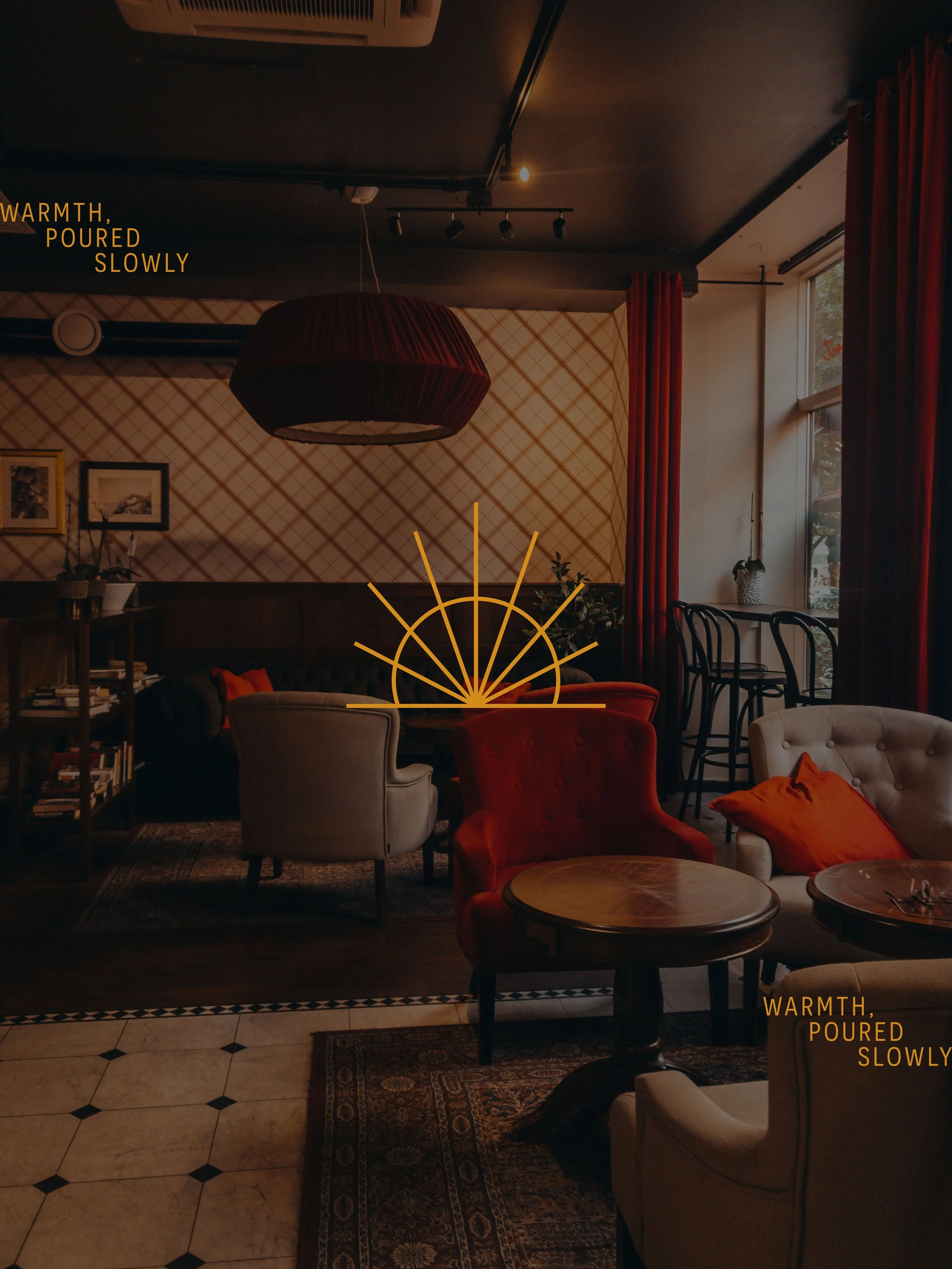 Cozy, warmly lit interior of a cafe or lounge with vintage furniture, including armchairs and small tables, decorated with cushions, plants, and framed artwork, with large windows draped with red curtains allowing natural light.