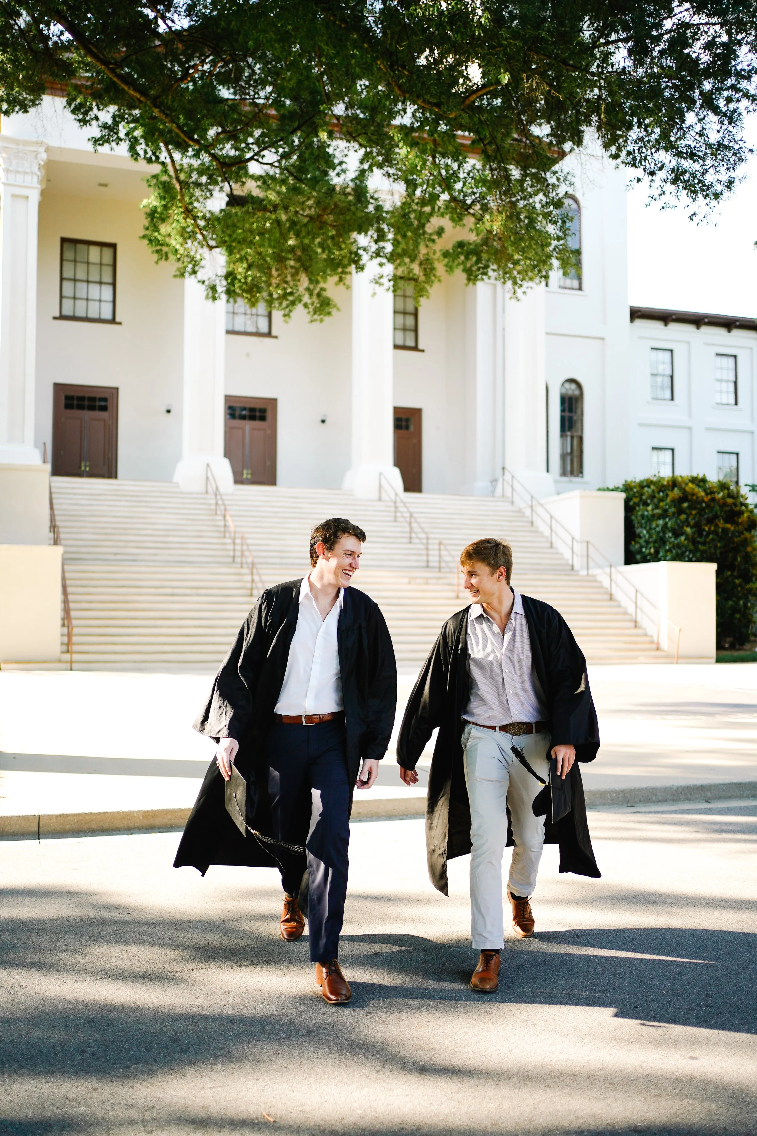 Two young men in graduation gowns walking and smiling in front of a large, white building with stairs and columns.