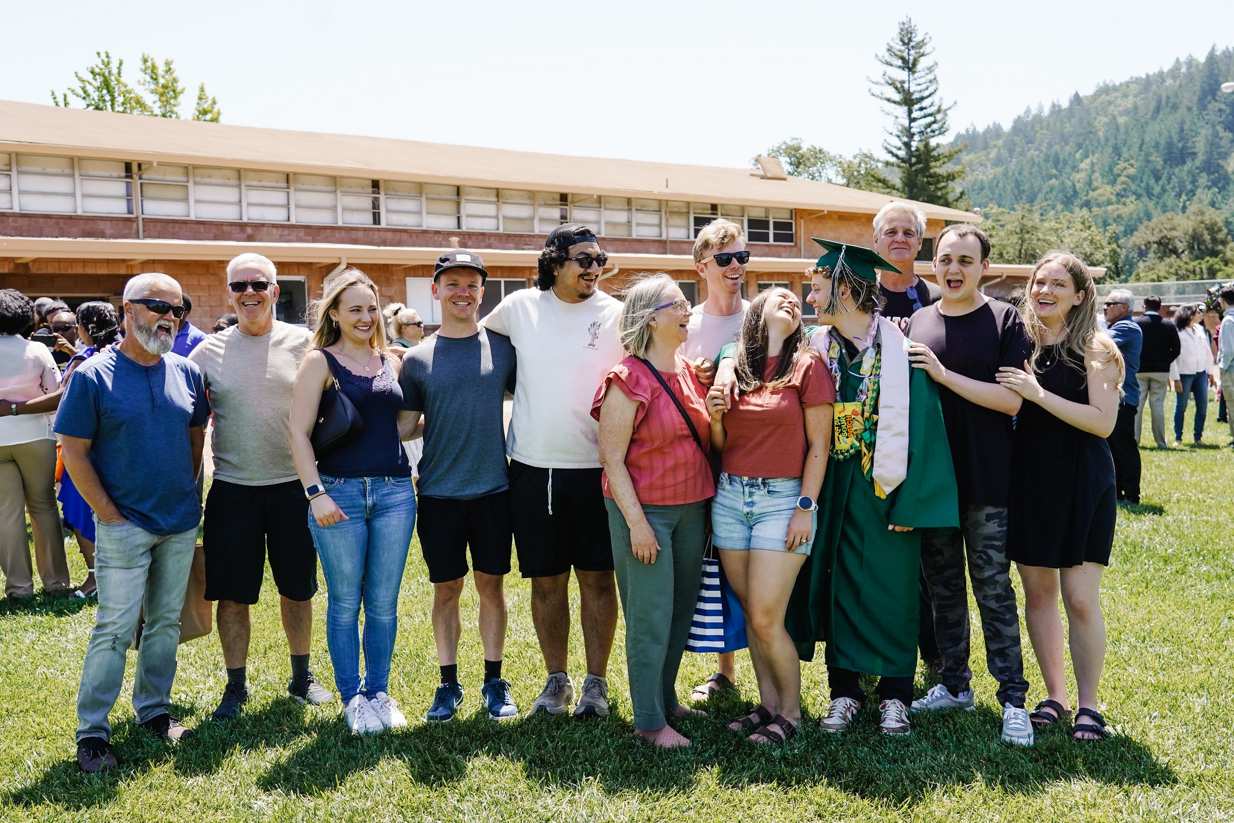 A group of diverse people celebrating a graduation outdoors on a grassy field, with a building and trees in the background.