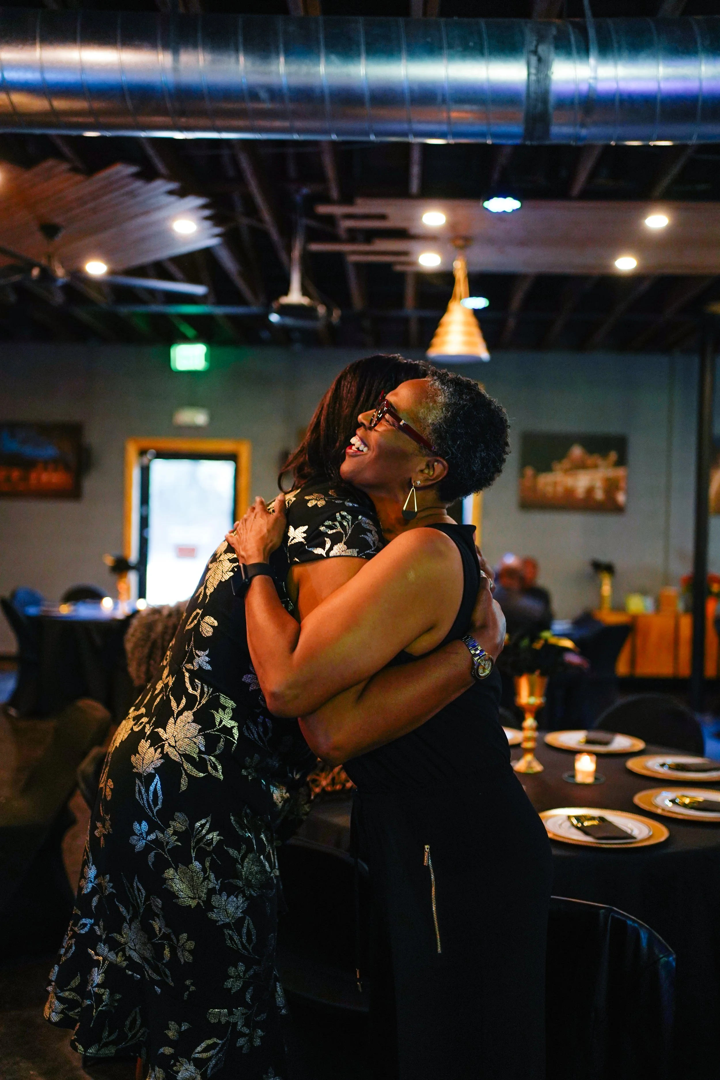Two women hugging each other at a dinner event in a dimly lit restaurant or banquet hall.