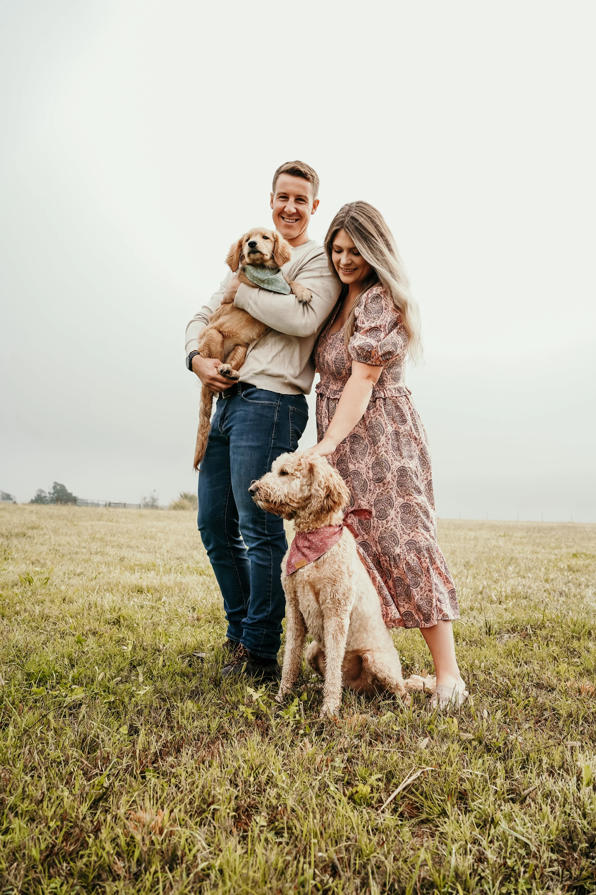A smiling couple with two dogs standing in a grassy field on a cloudy day.