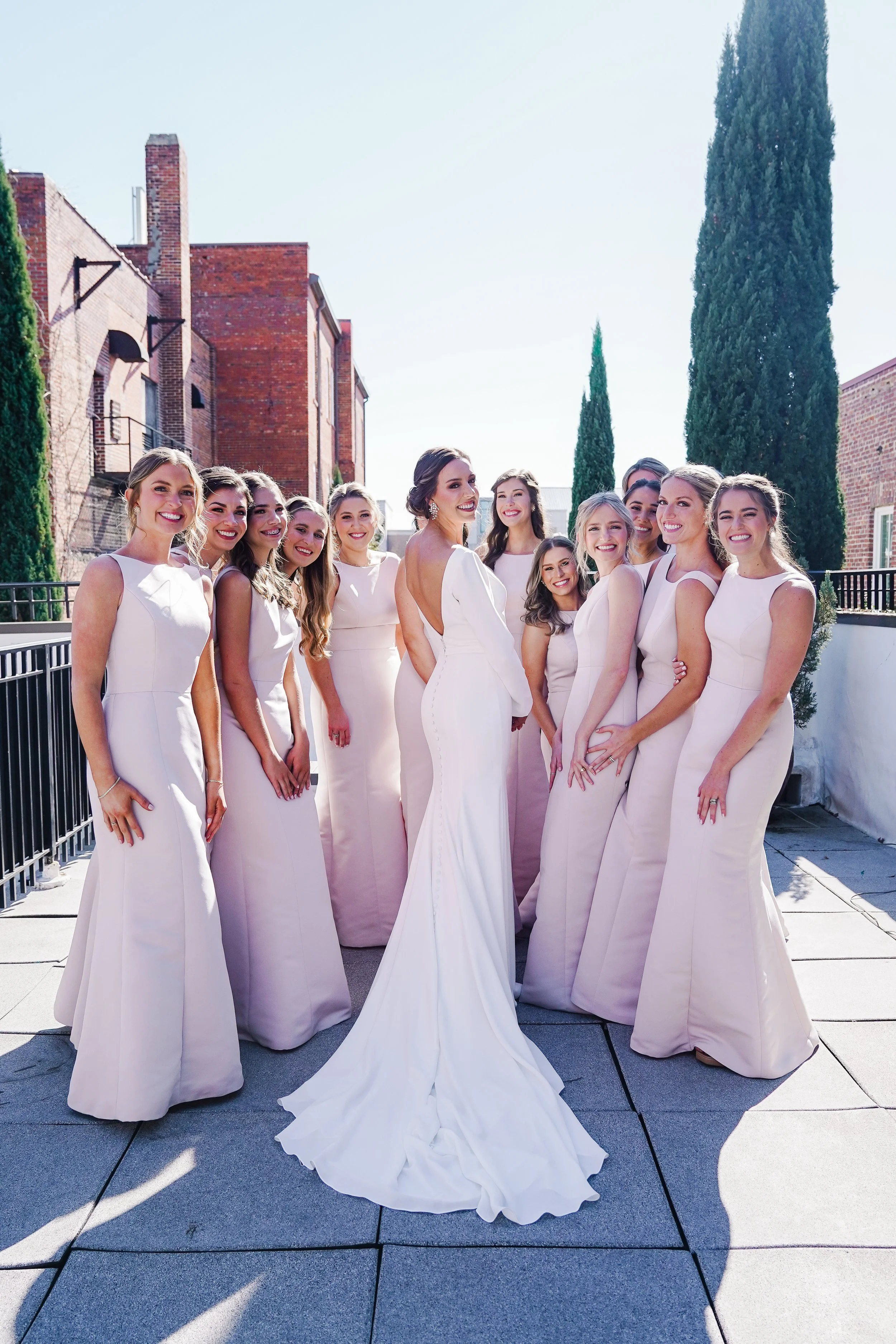 A bride in a white wedding dress surrounded by her bridesmaids in matching pale pink dresses outdoors on a sunny day with brick buildings and tall trees in the background.