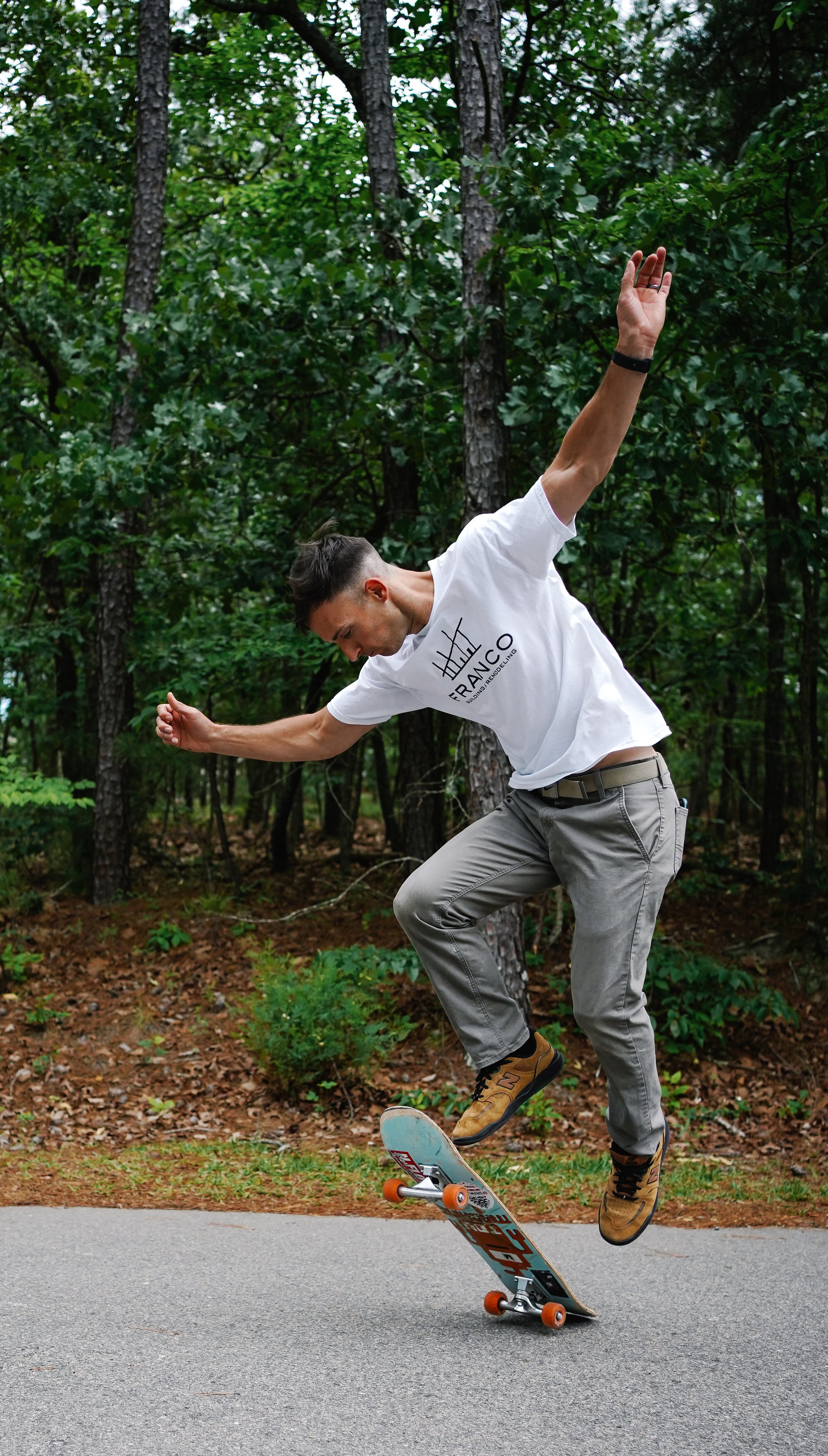 A man balancing on a skateboard outdoors on a paved path in a wooded area, wearing a white T-shirt, beige pants, and brown shoes.