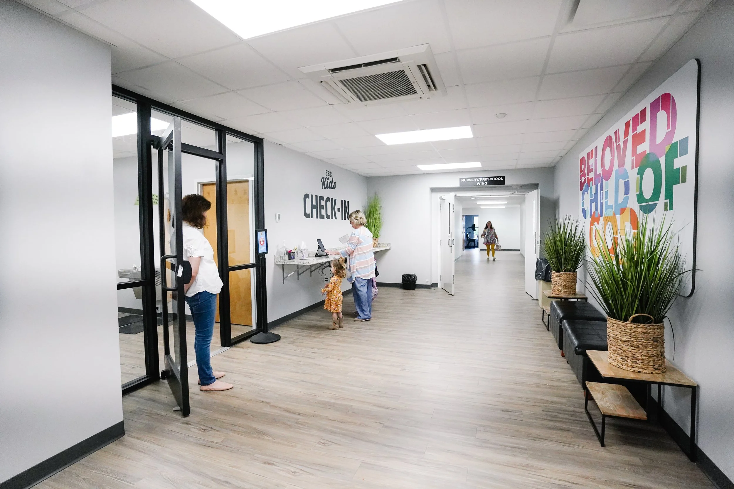 Interior of a children's healthcare or educational facility showing a check-in area on the left, a hallway with a colorful sign on the right, and two women with children near the check-in desk, with one woman waiting and the other interacting with a 