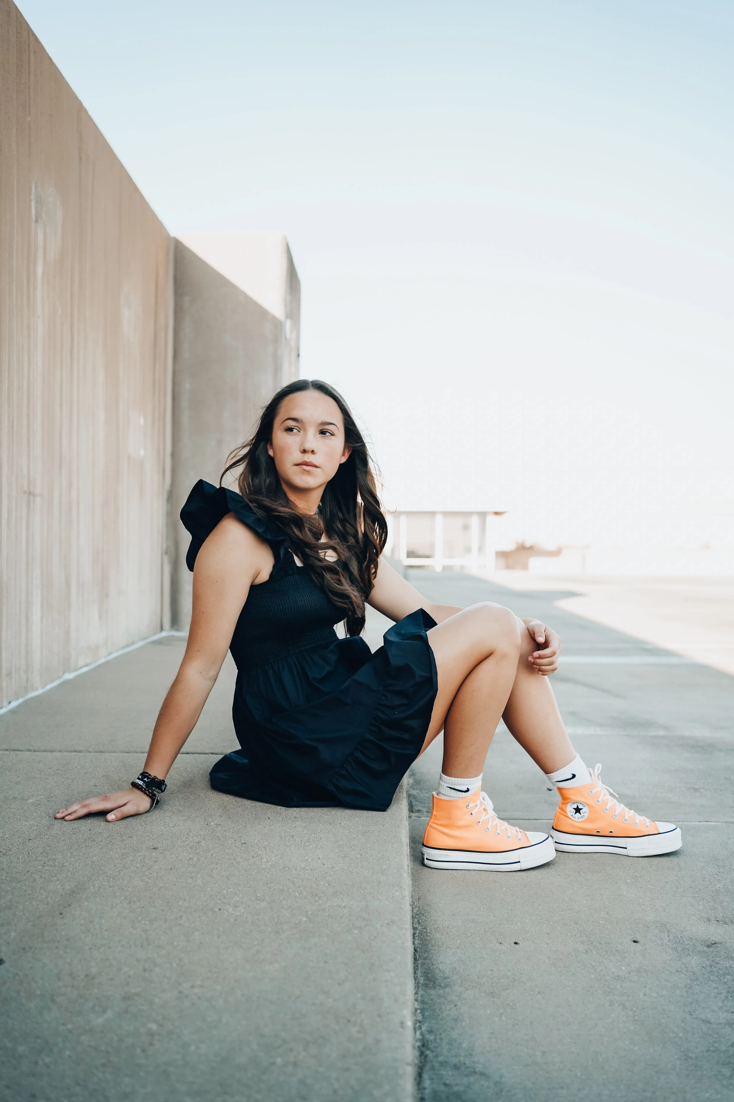 A young woman in a black dress and orange sneakers sitting on the sidewalk next to a concrete wall, with a clear sky background.