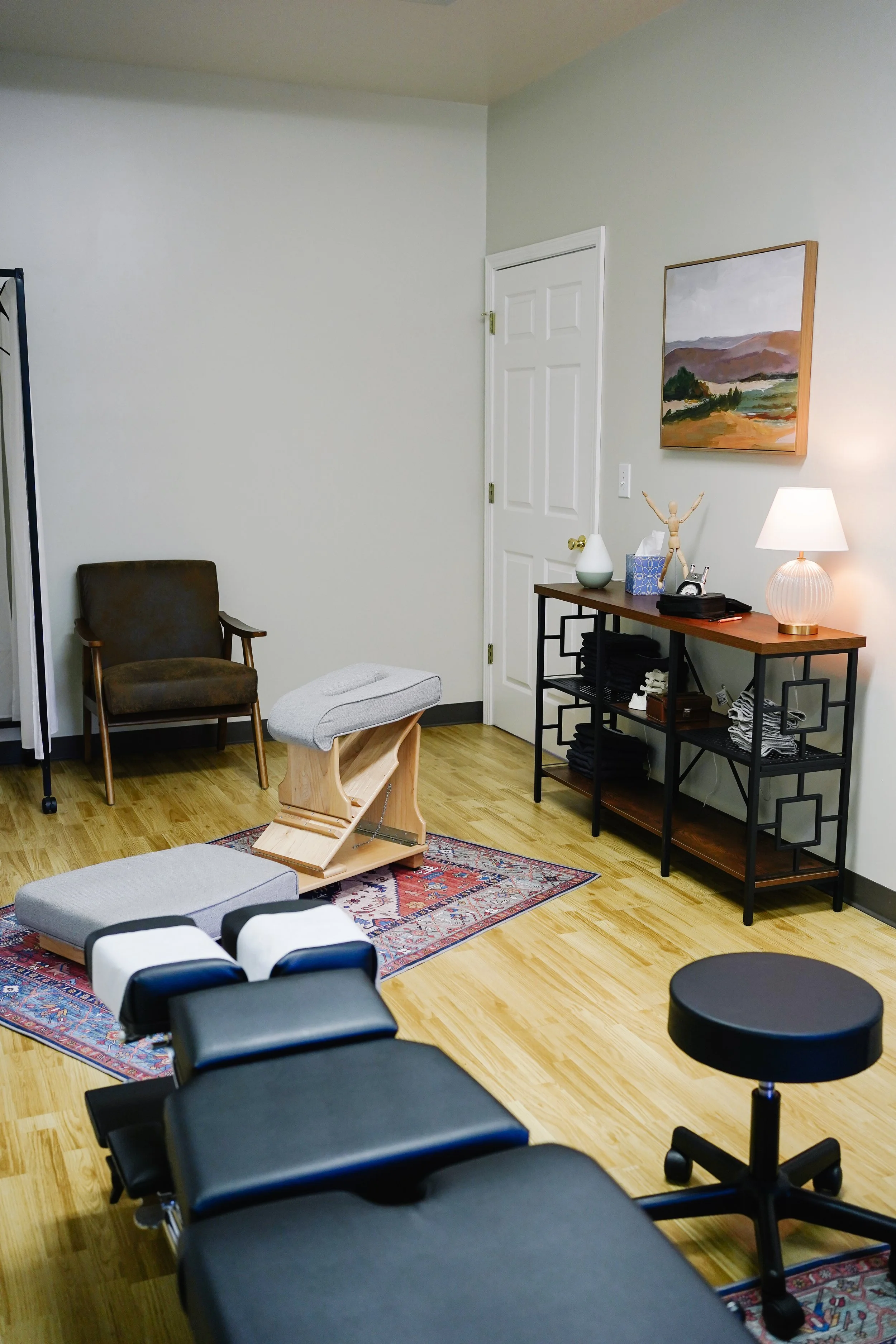 A room with a treatment table, a brown chair, a small rug, and a cart with folded towels and a lamp, suggesting a physical therapy or medical office.