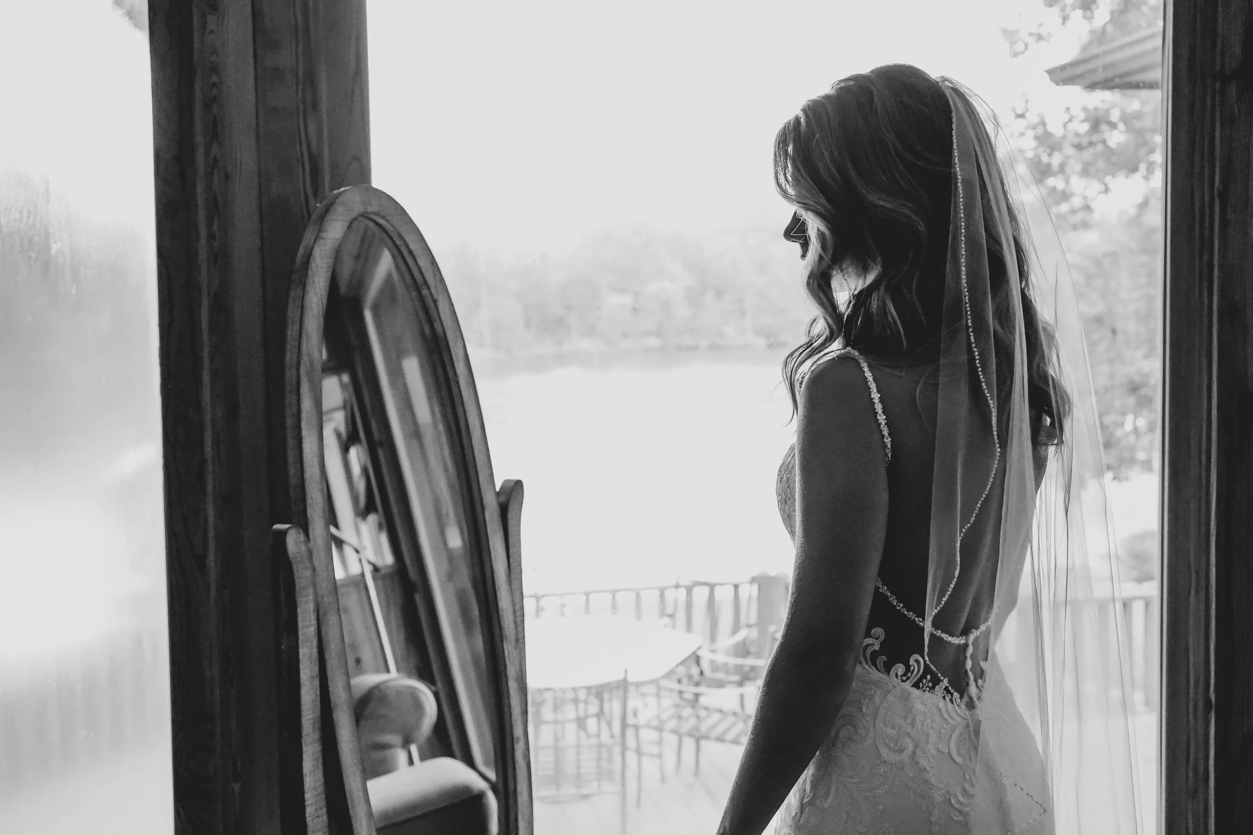 A bride standing indoors by a mirror, looking outside through a door, wearing a wedding dress and veil, with natural light illuminating her silhouette.