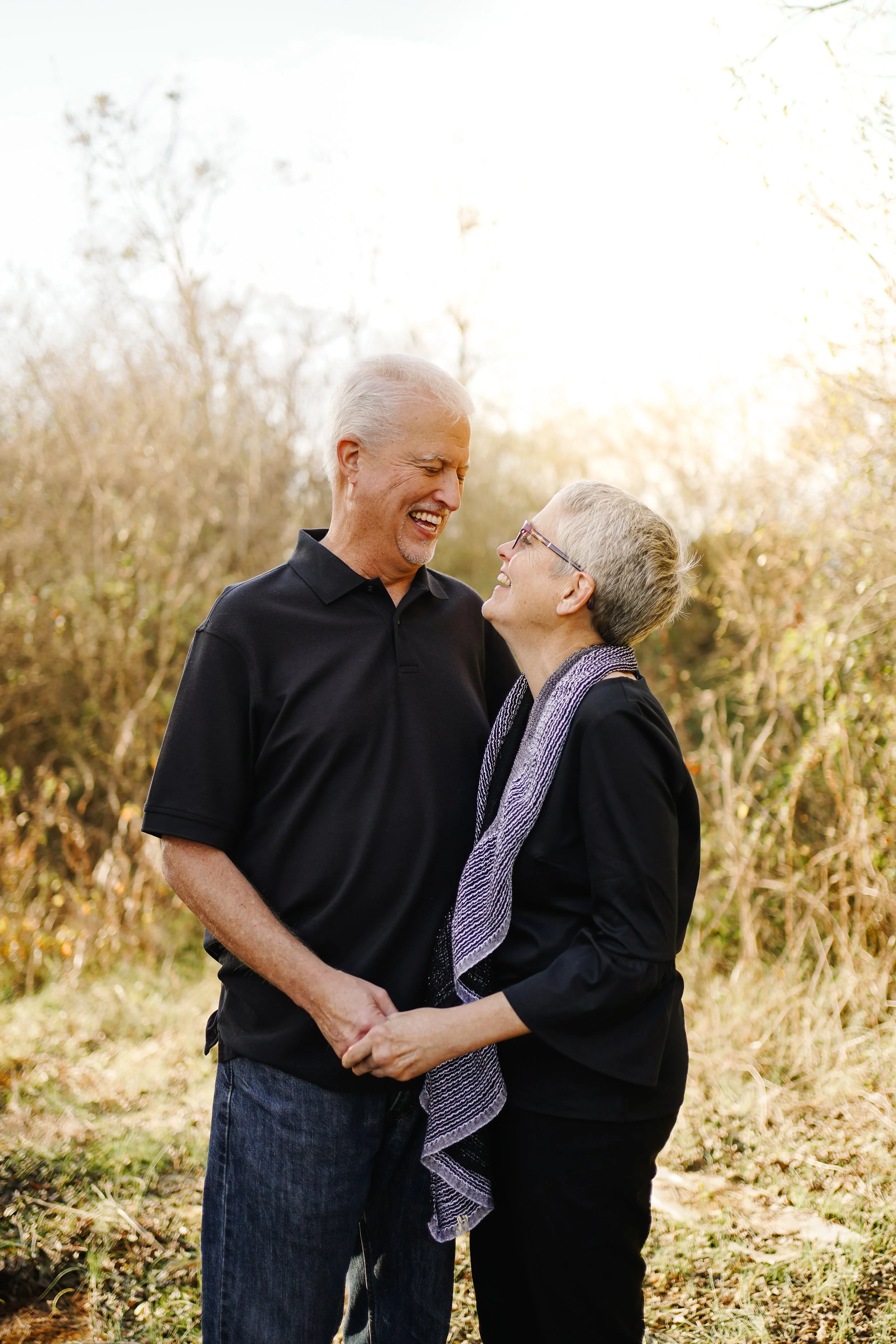 An elderly couple smiling and holding hands outdoors during daytime, surrounded by trees with fall foliage.