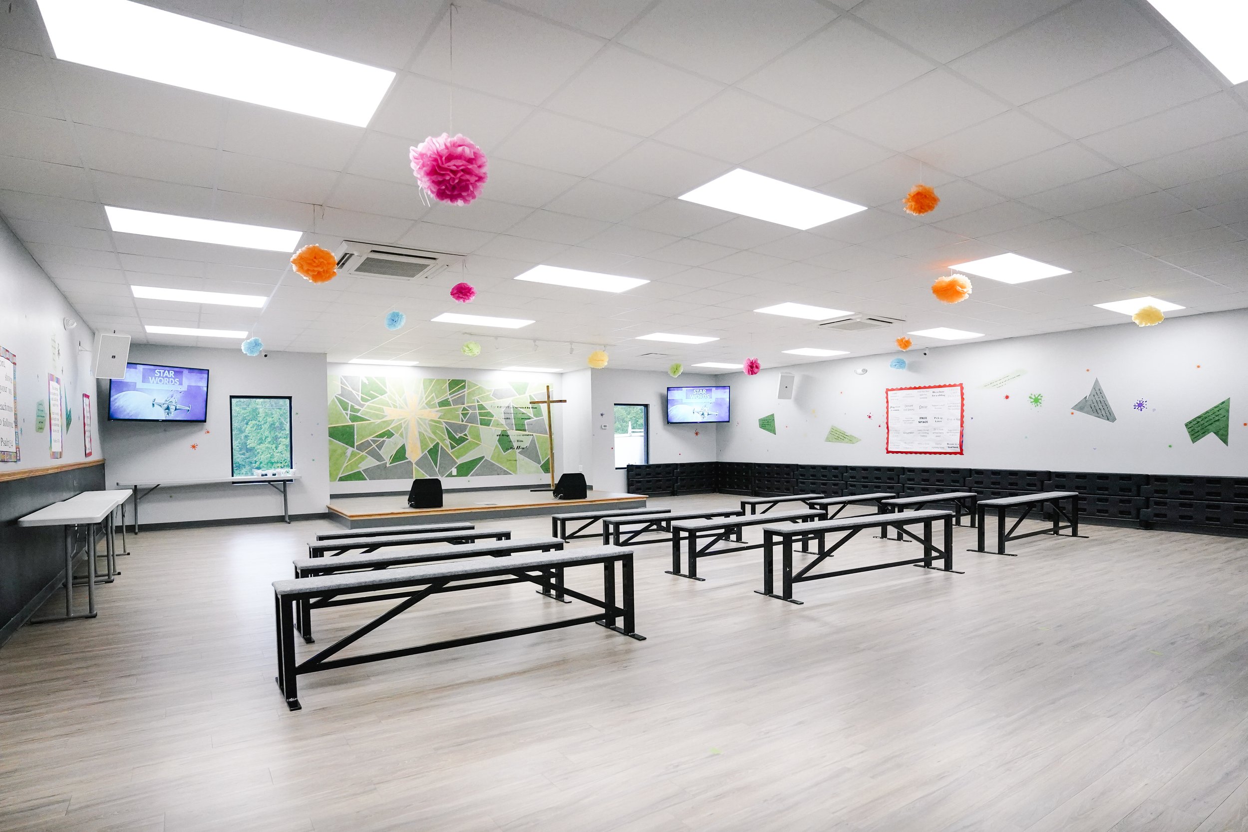 Empty church or community hall with a stage, colorful hanging paper decorations, benches, and wall murals.