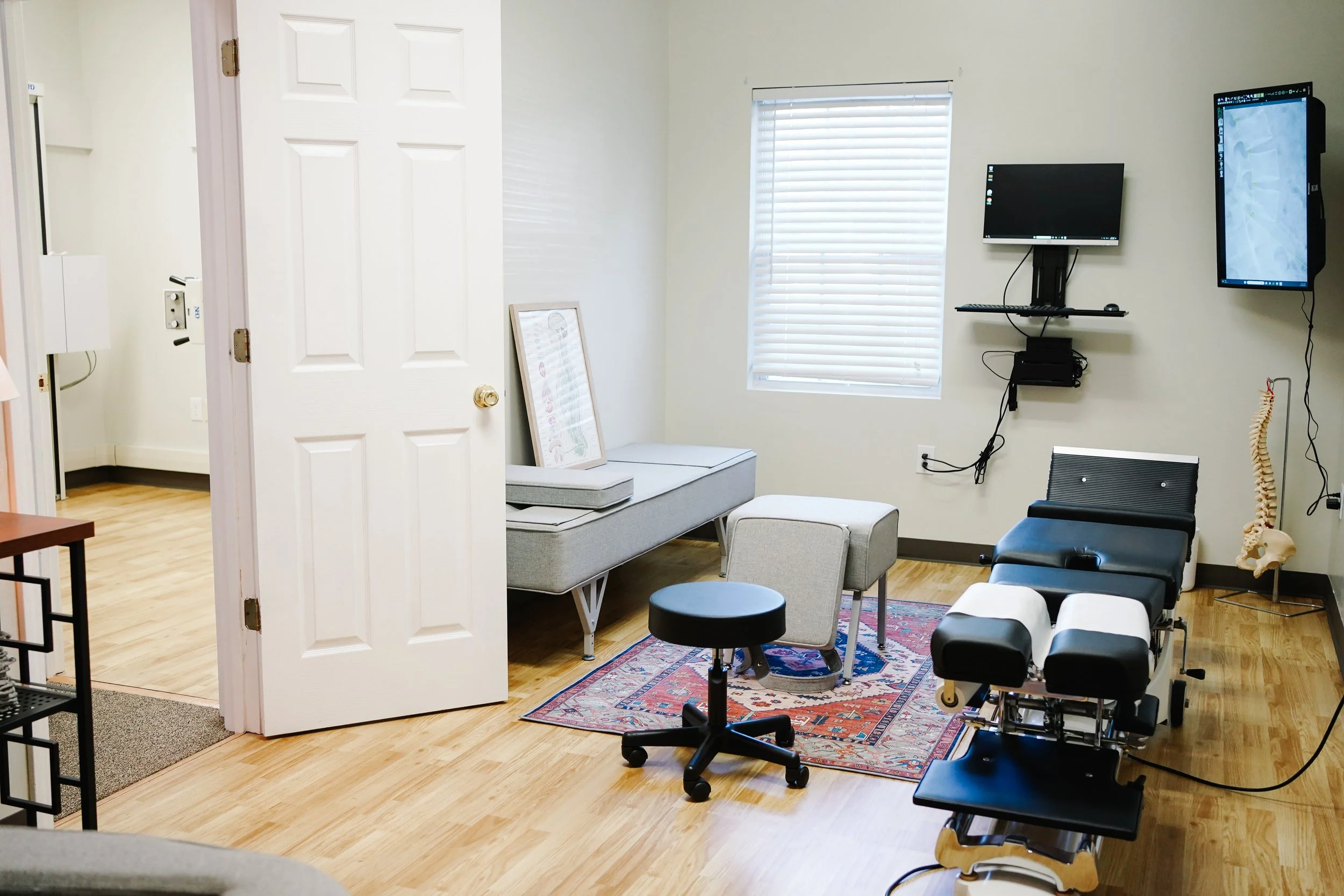 Medical consultation room with examination table, desk, chairs, wall-mounted monitors, and medical equipment.