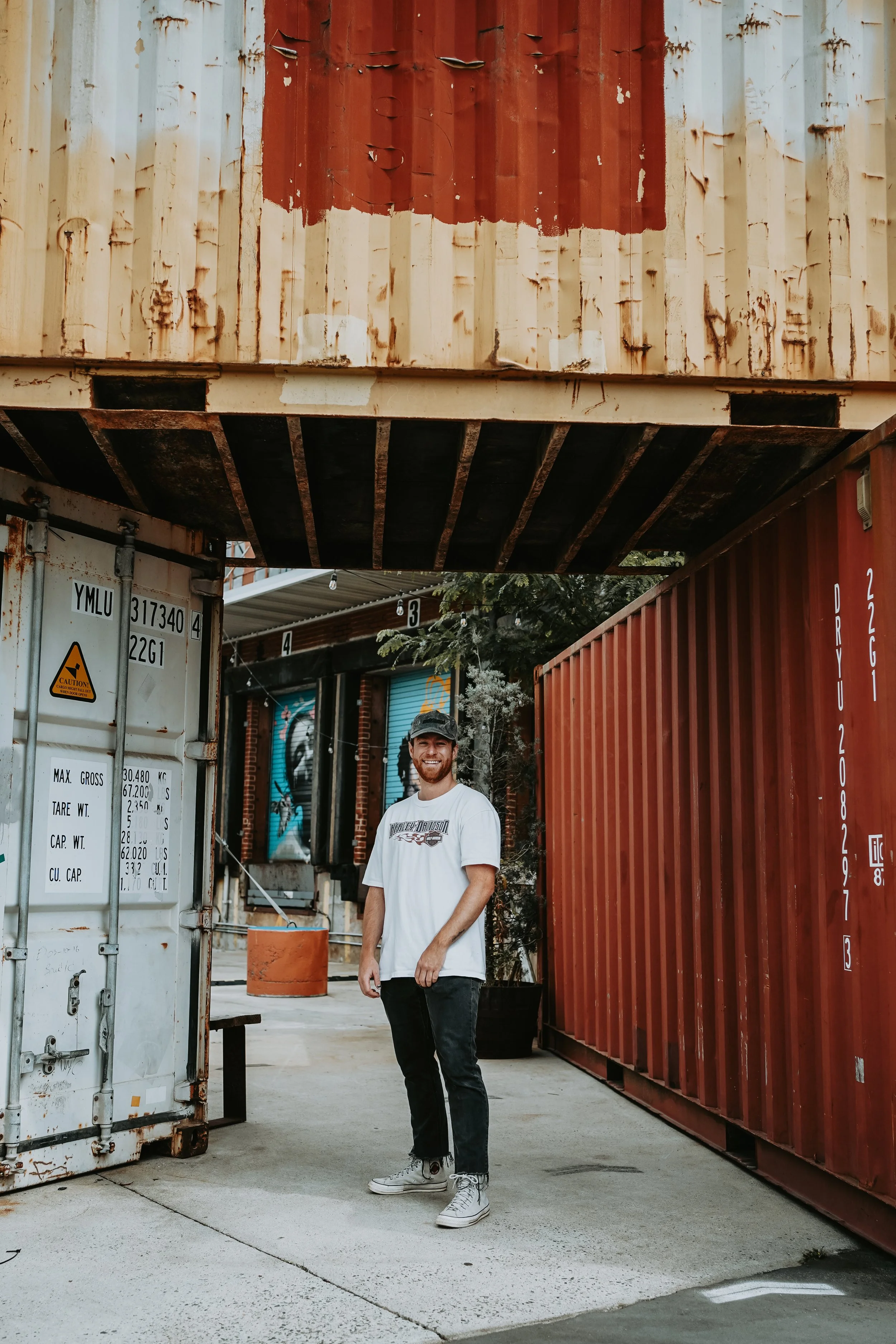 A man with a beard, wearing a gray cap, white t-shirt, black pants, and gray sneakers, stands smiling on a sidewalk under a large shipping container and next to another container. There are graffiti murals on the building behind him.