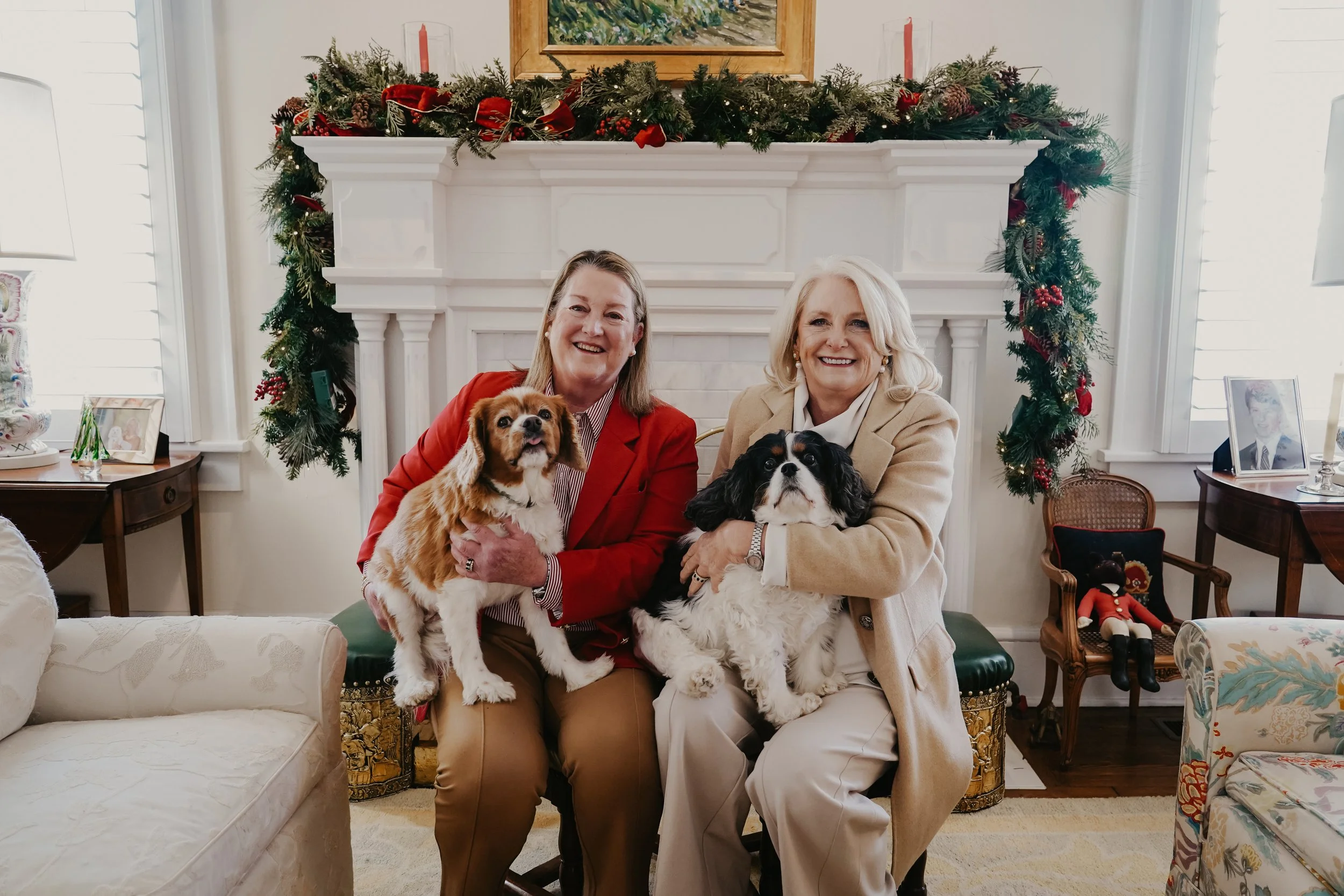 Two women sitting in a living room with Christmas decorations, each holding a dog. One woman is wearing a red blazer and the other a beige blazer. They are smiling and sitting in front of a decorated fireplace mantel.