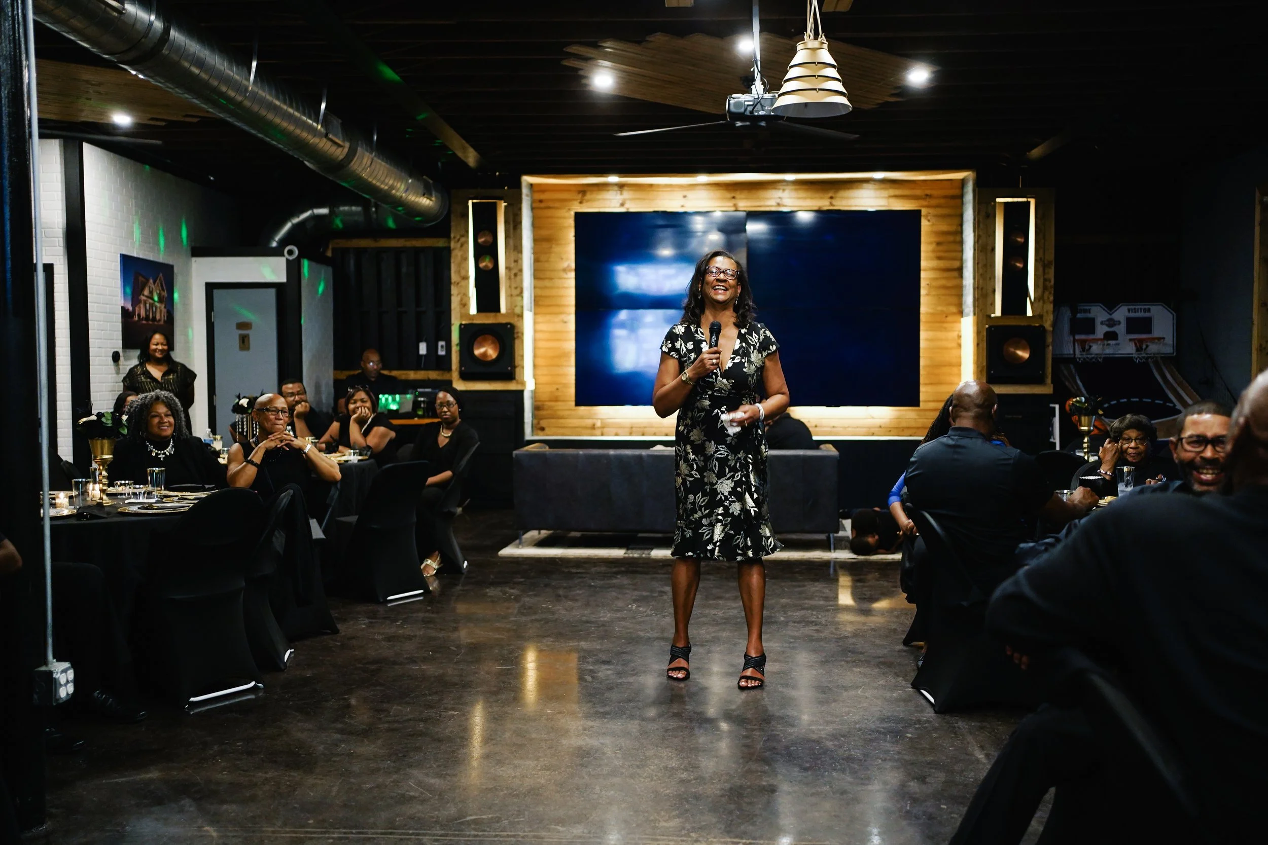 A woman in a floral dress and heels speaking into a microphone at a gathering in a dimly lit venue with round tables and guests watching her.