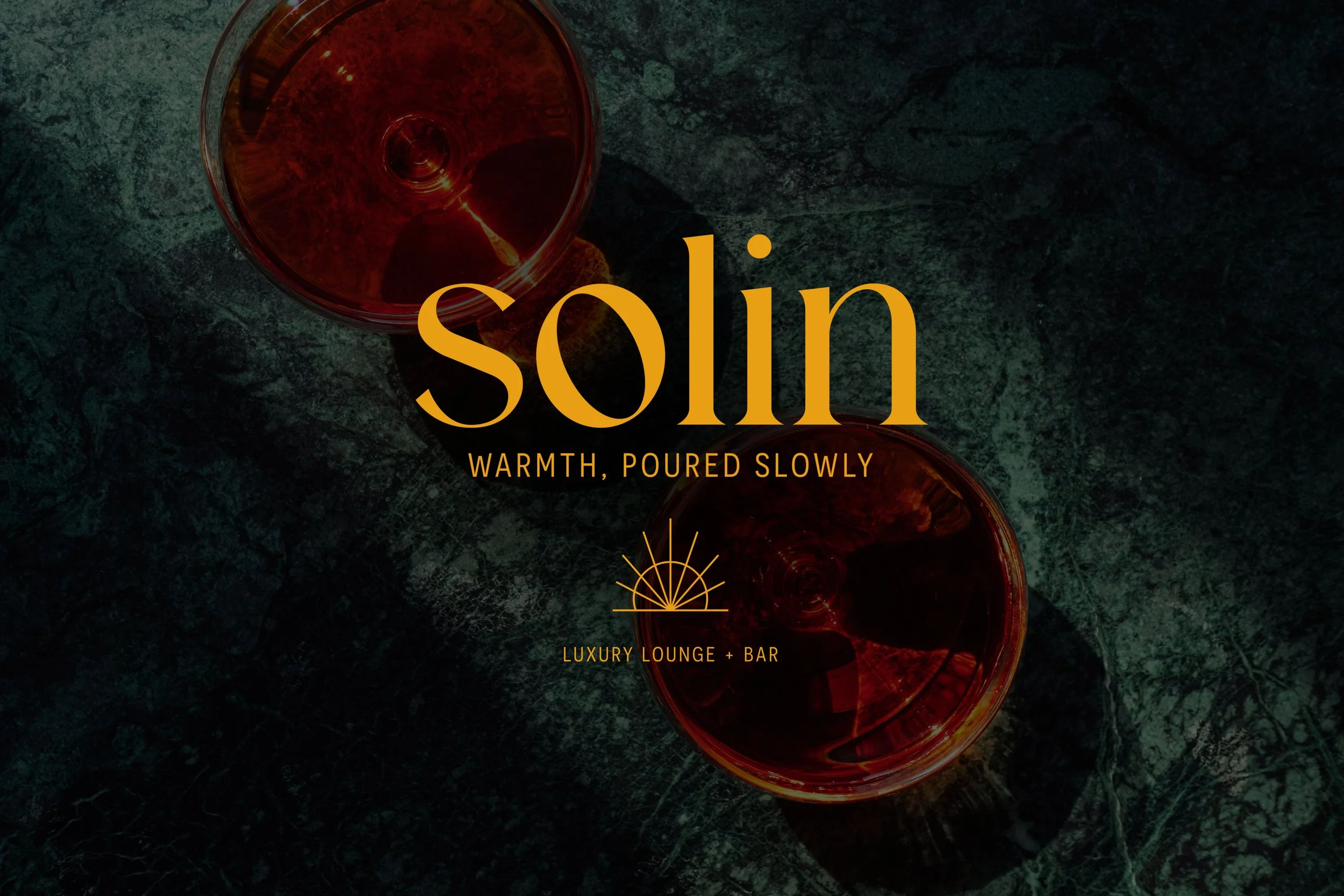 Two glasses of red wine on a dark green marble surface with the word 'solin' in large orange letters, and below it the phrase 'warmth, poured slowly' and an icon of a rising sun, followed by the words 'luxury lounge + bar'.