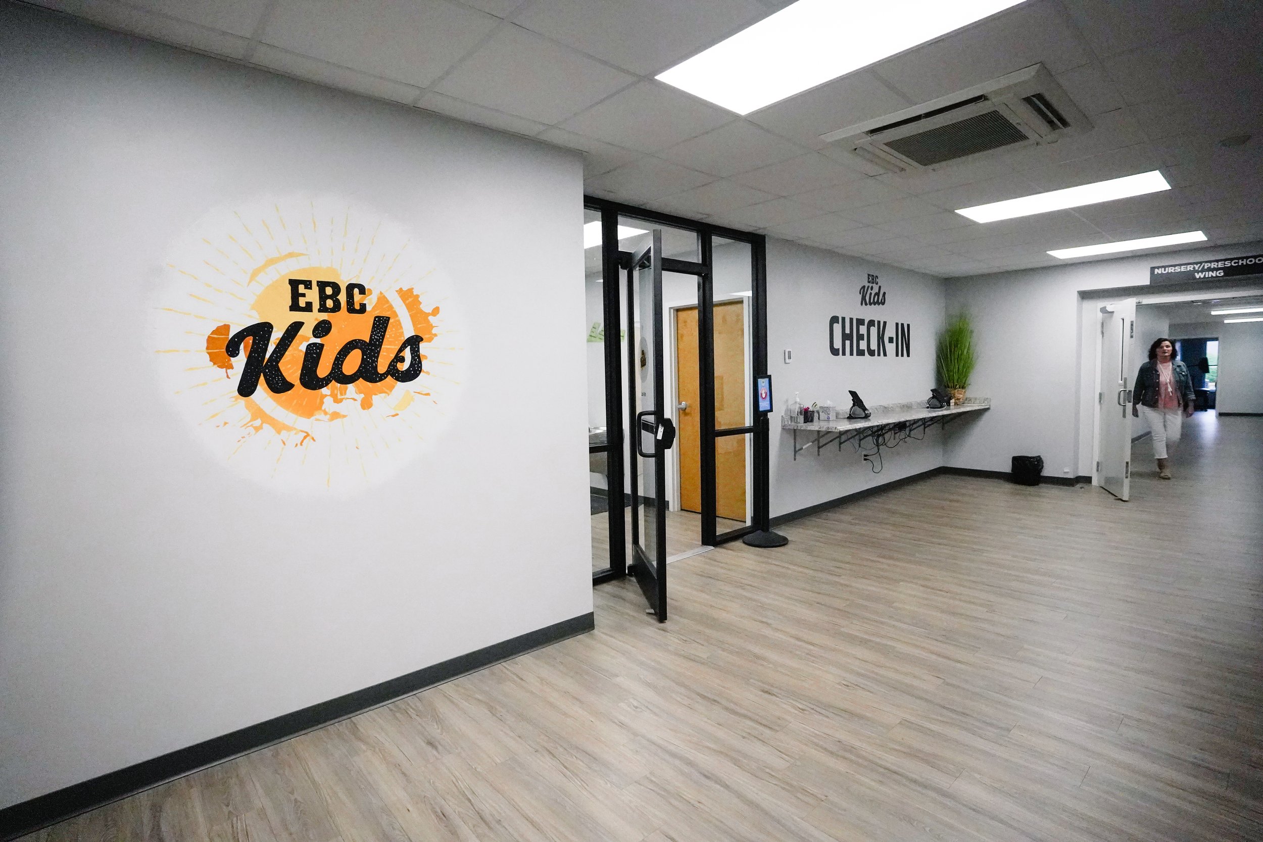 Interior of a children's program check-in area at EBC Kids, with a wall sign, a desk, and a woman walking in the background.