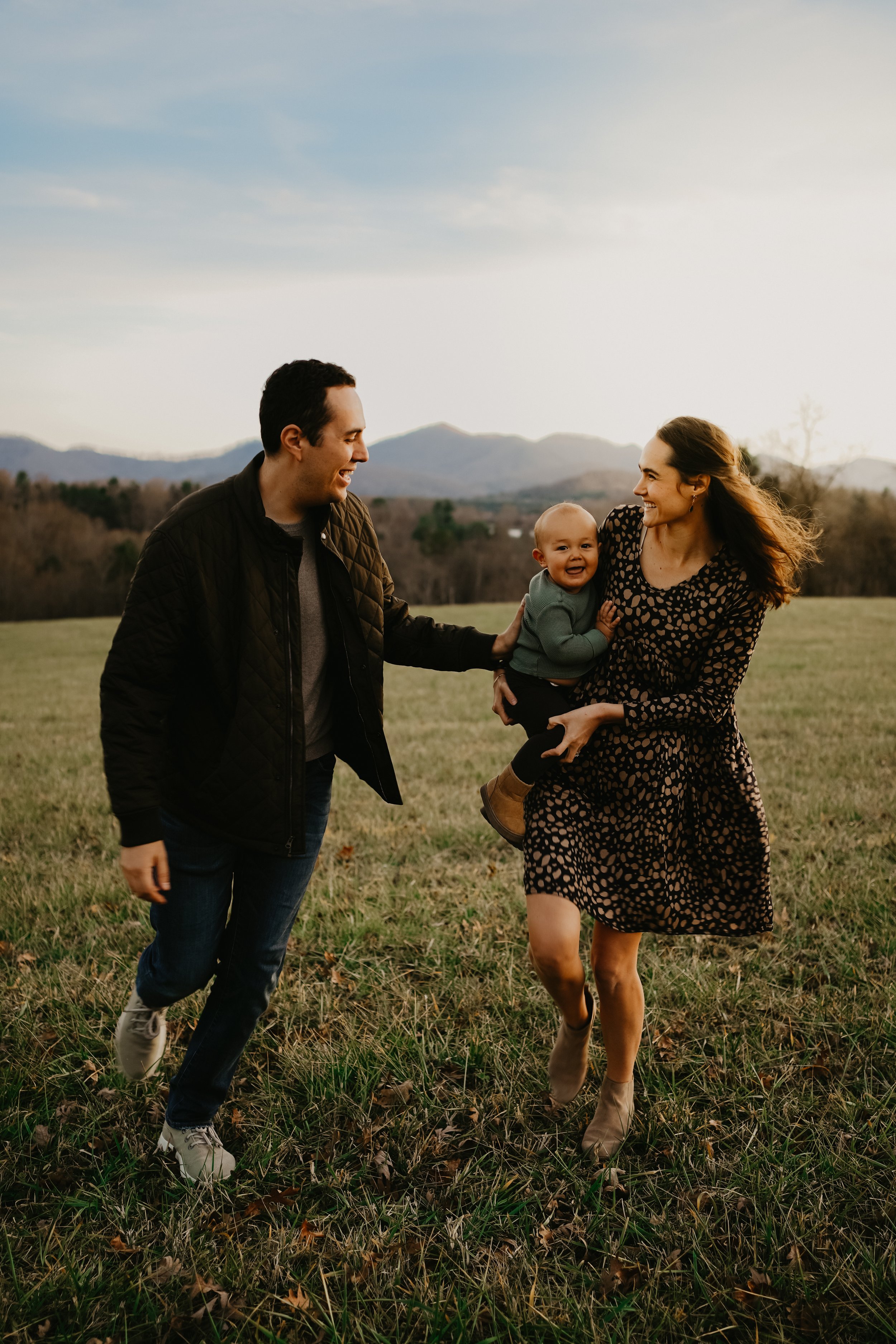 A family of three running through a grassy field at sunset, with mountains in the distance. The woman is holding a smiling child, and a man is reaching out to her.