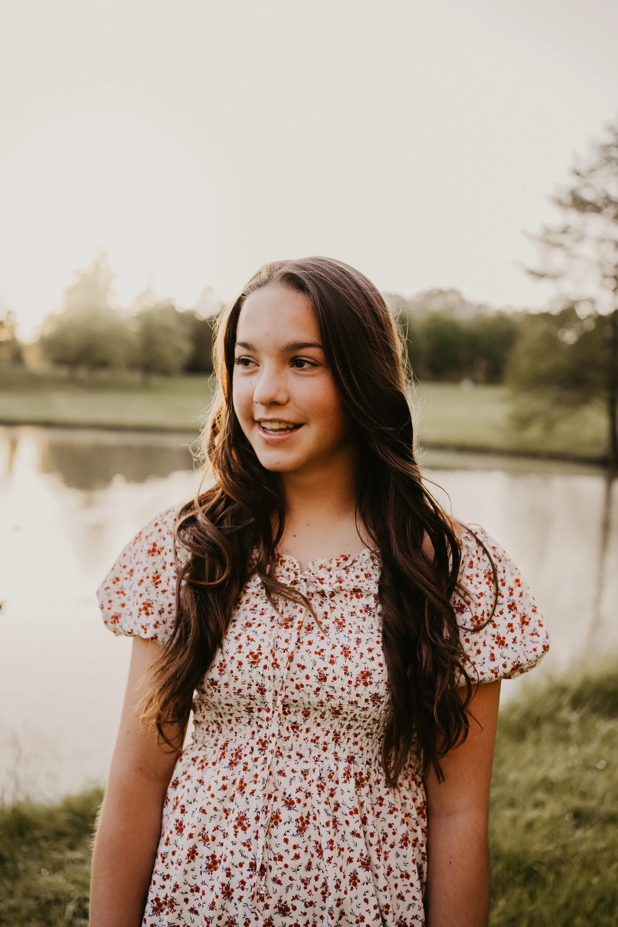 A young woman with long dark hair smiling outdoors near a pond during sunset.
