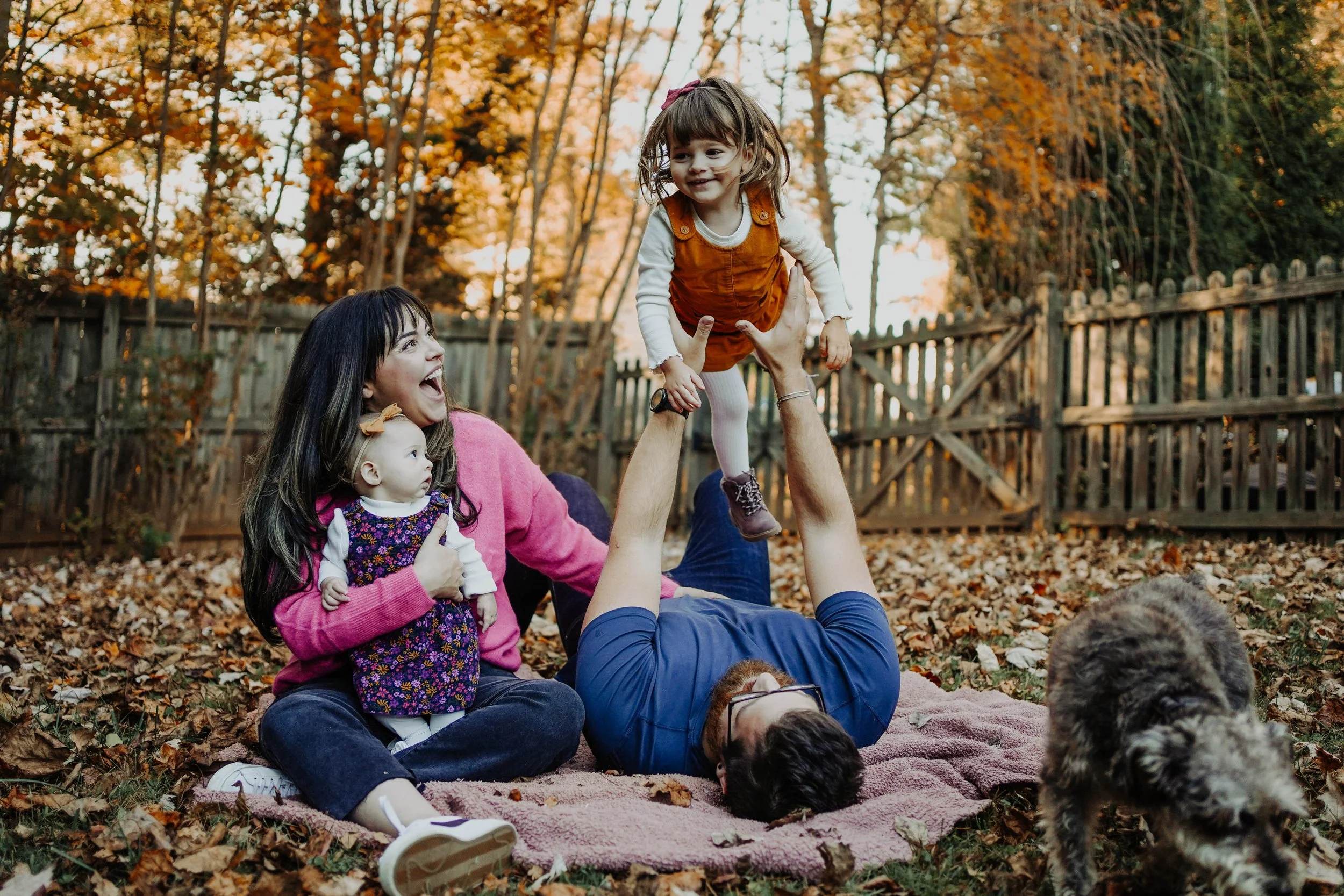 Family playing outdoors in fall, with autumn leaves on the ground and trees, a woman holding a baby, a man laying on a blanket lifting a young girl, and a dog nearby.