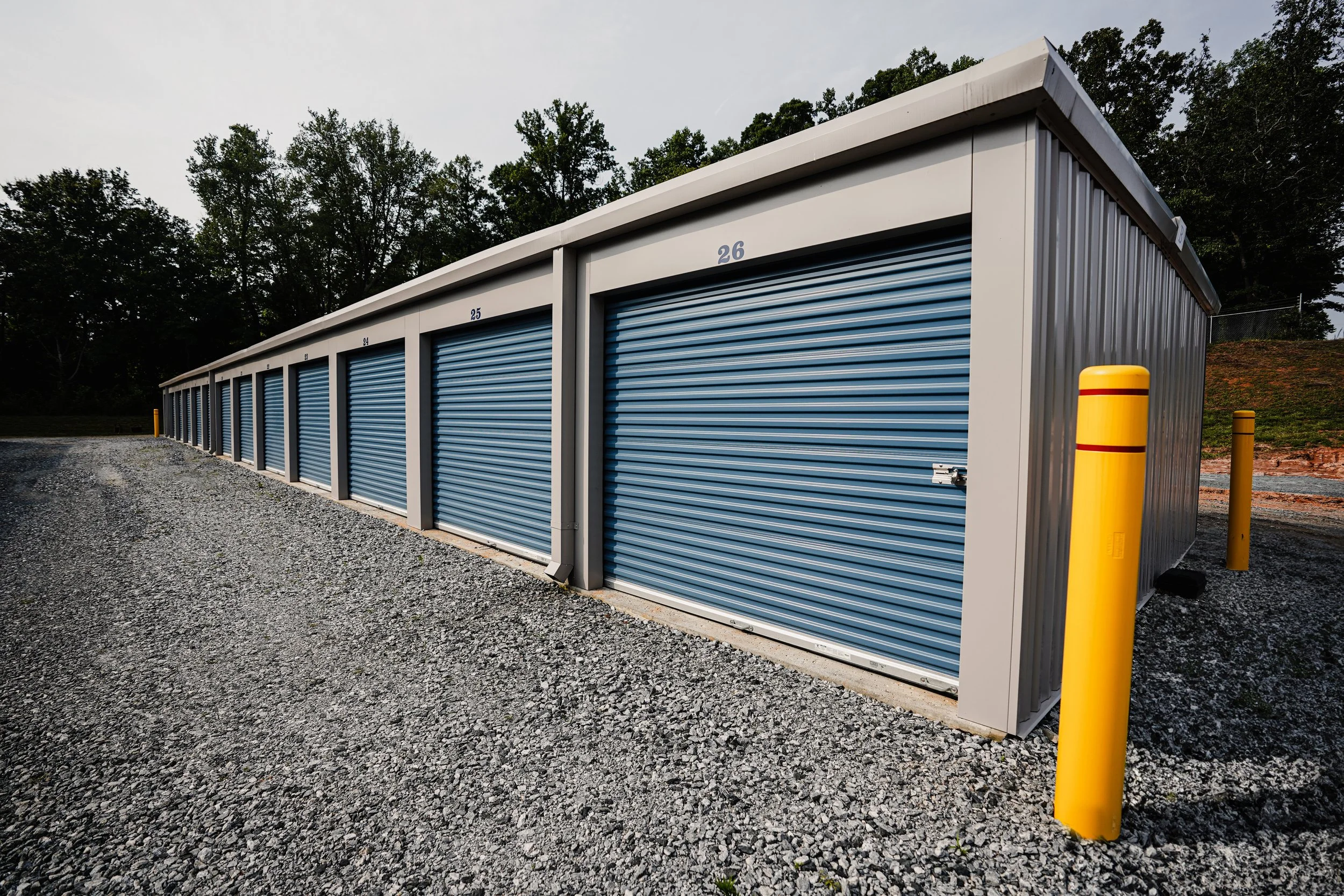 A row of blue storage unit doors with numbers, set under a white metal roof, with yellow protective bollards in front and background of trees.