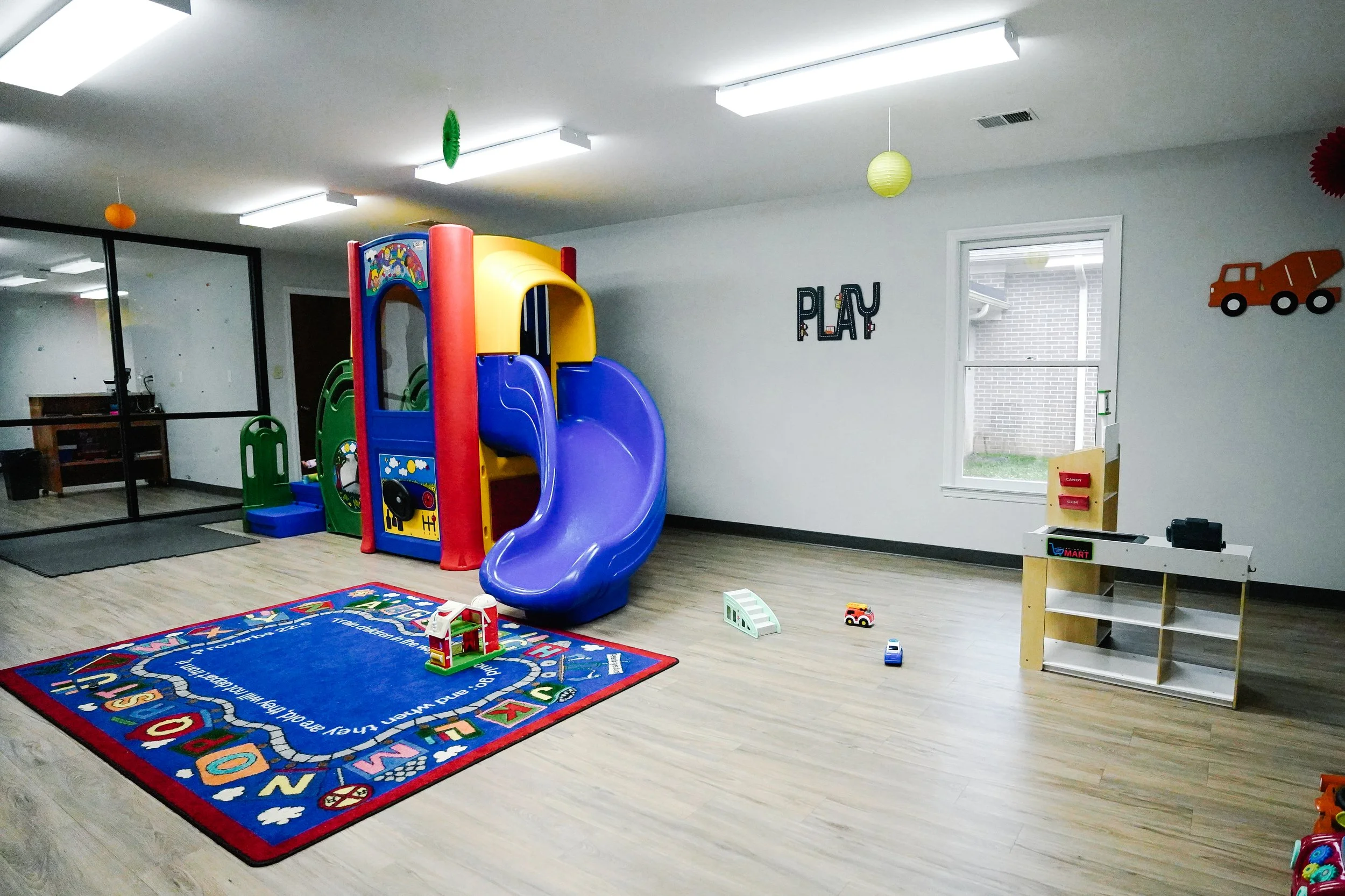 An empty indoor children's playroom with a colorful plastic playground slide, a play mat with alphabet and city designs, small toy vehicles, and wall decorations including a cage, a truck, and a bicycle, along with a window and a glass partition.