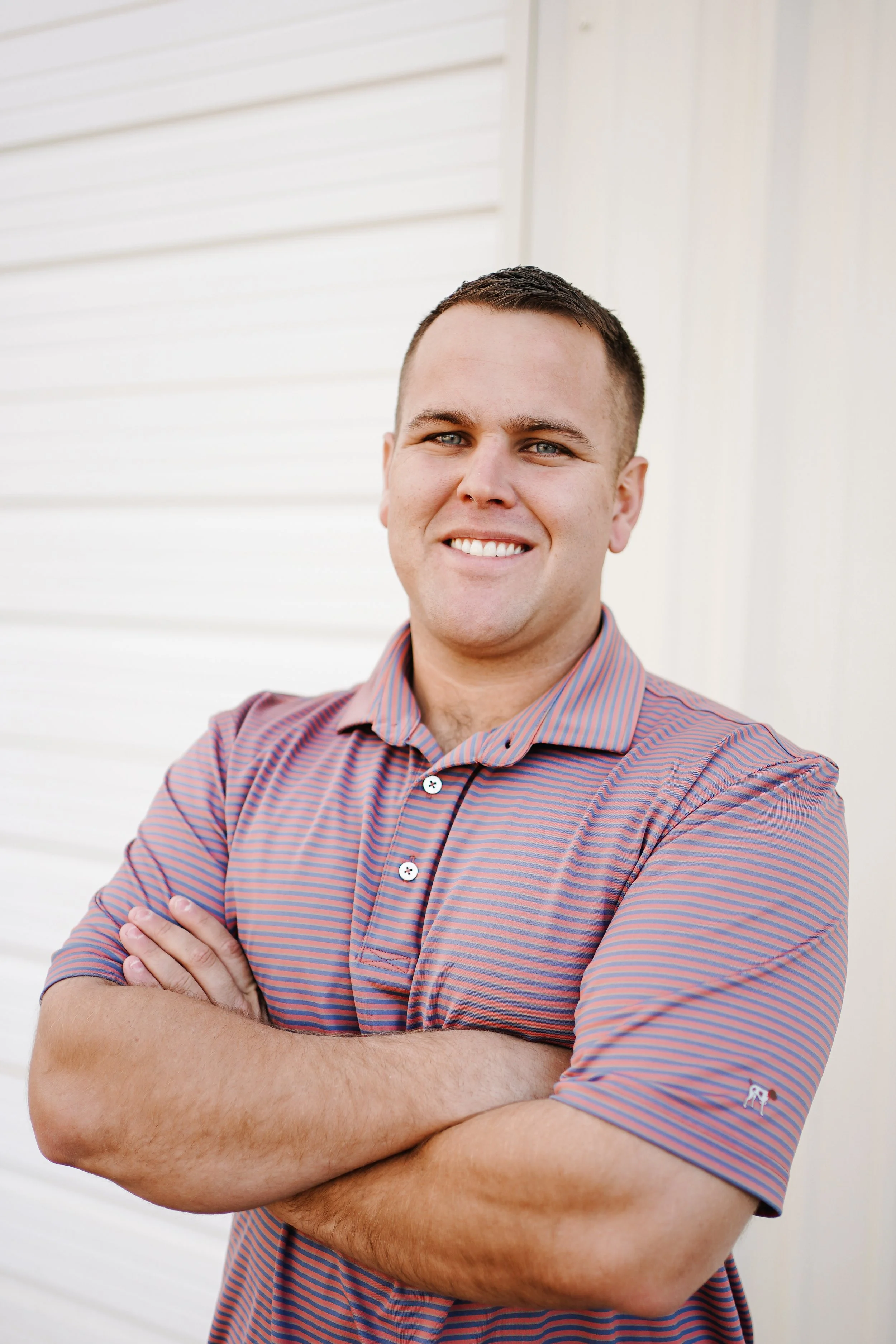 A smiling man with short dark hair standing outside with arms crossed, wearing a red and blue striped polo shirt, in front of a white building with horizontal siding.