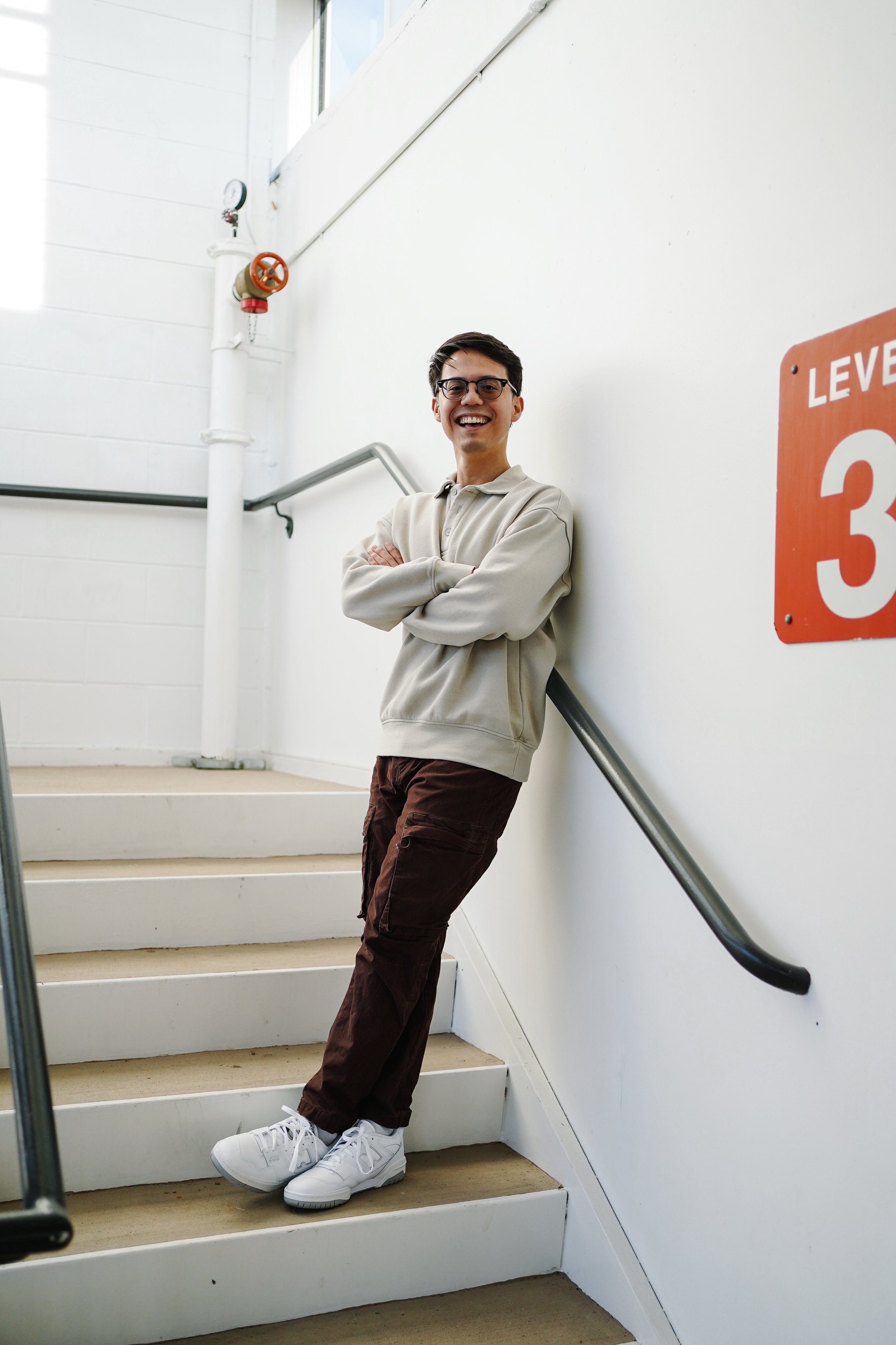 A smiling young man with glasses and dark hair, standing on a staircase with arms crossed, leaning against a white wall in an industrial building, wearing a beige hoodie, brown cargo pants, and white sneakers.