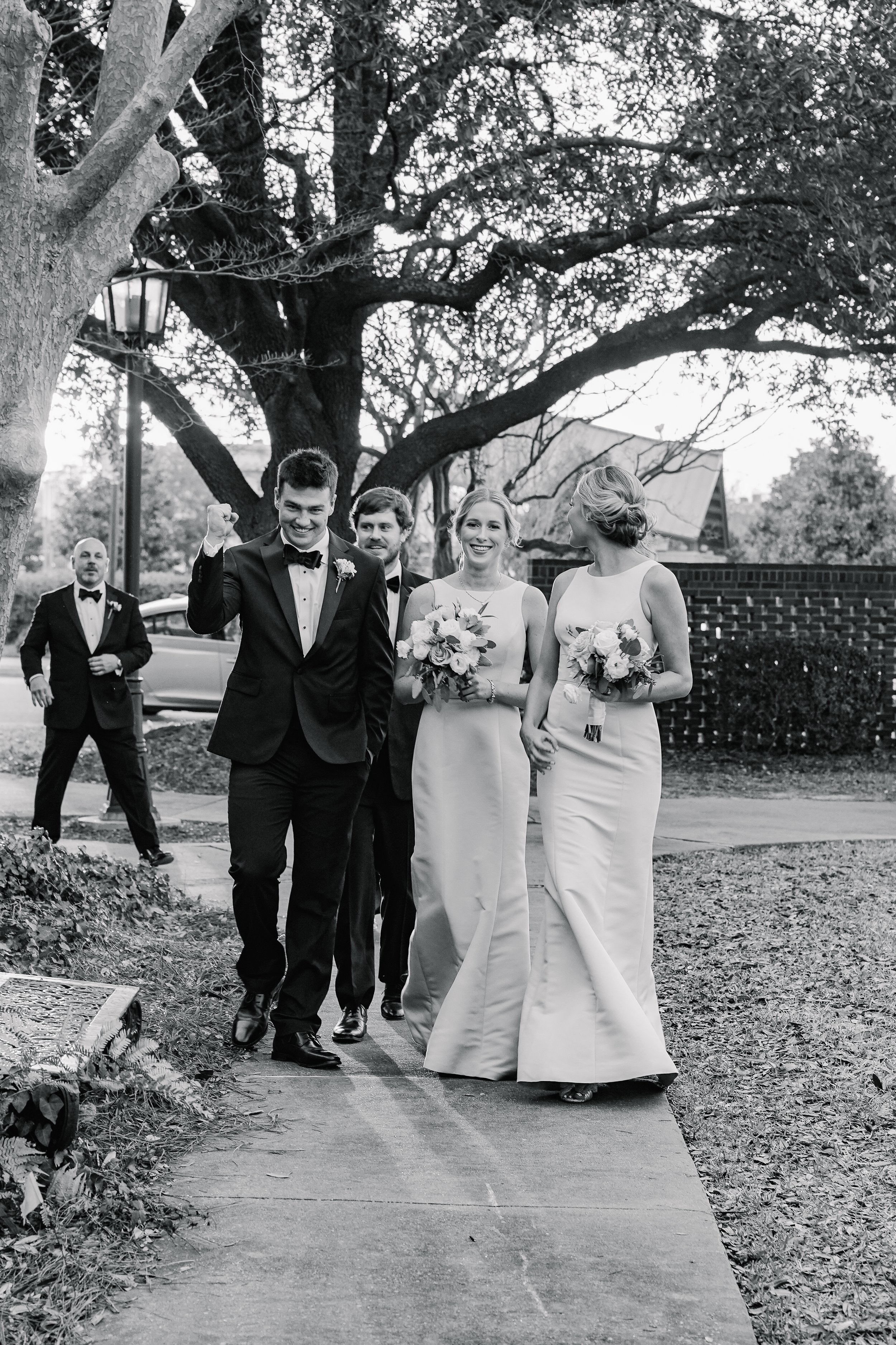 Black and white photo of a wedding procession with a bride, groom, and bridesmaids walking arm in arm outdoors under a large tree, with two men in tuxedos following in the background.