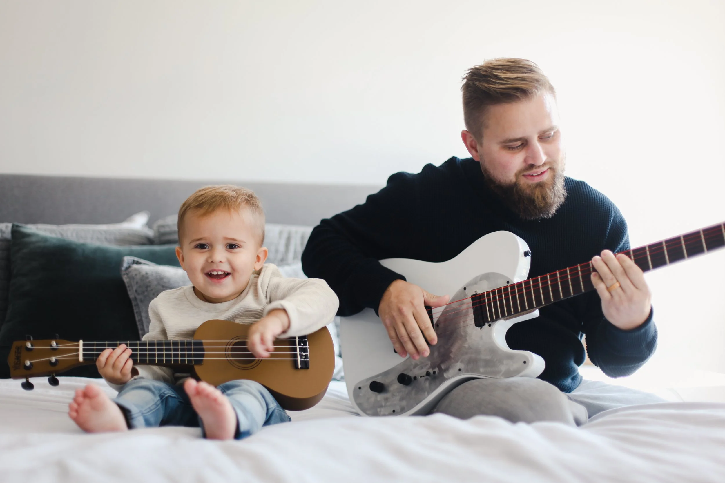 A young man and a small boy sitting on a bed playing guitars, with pillows and a neutral-colored headboard in the background.