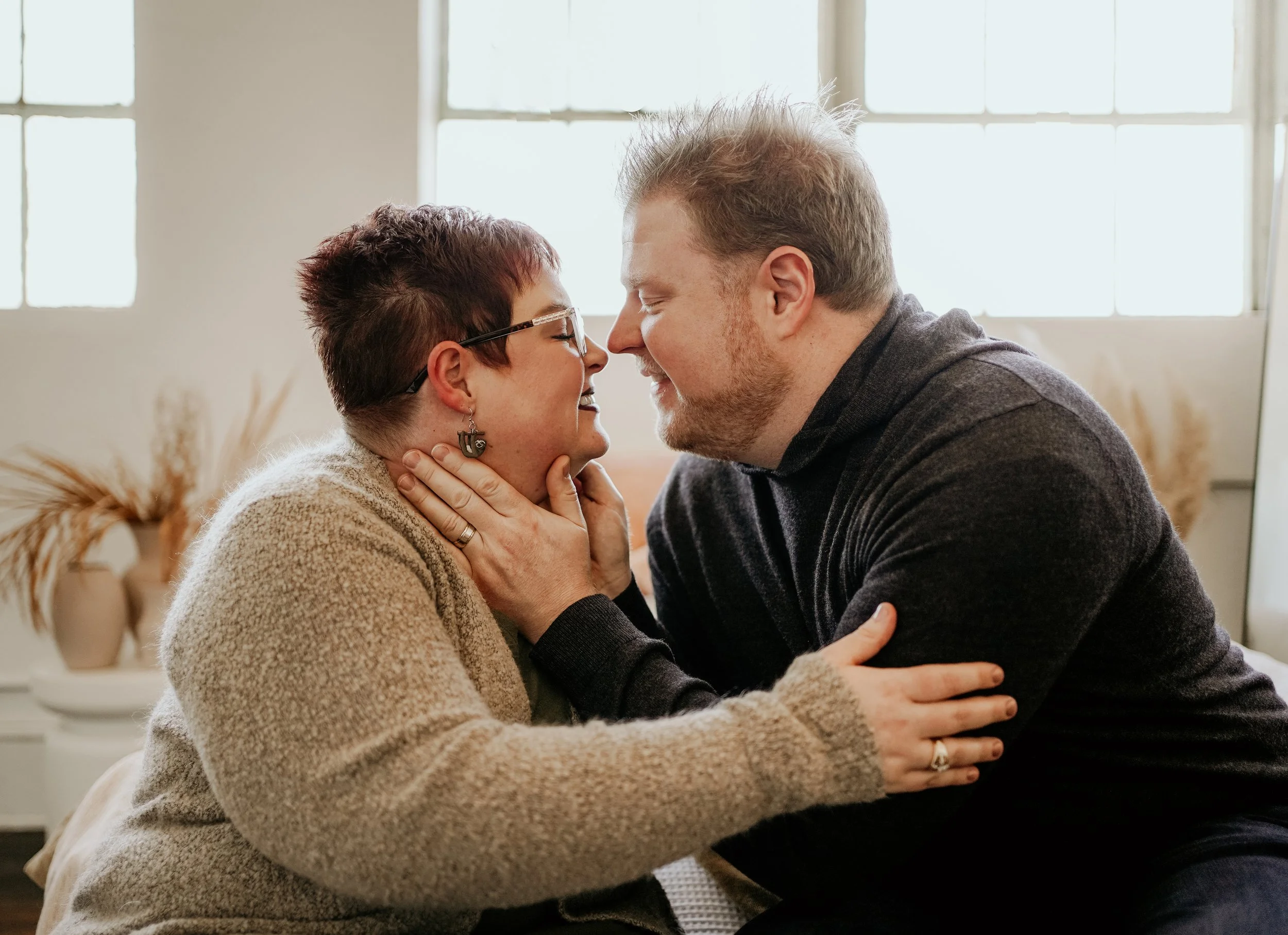 A couple touching foreheads and smiling in a warmly lit room with large windows and decorative plants in the background.
