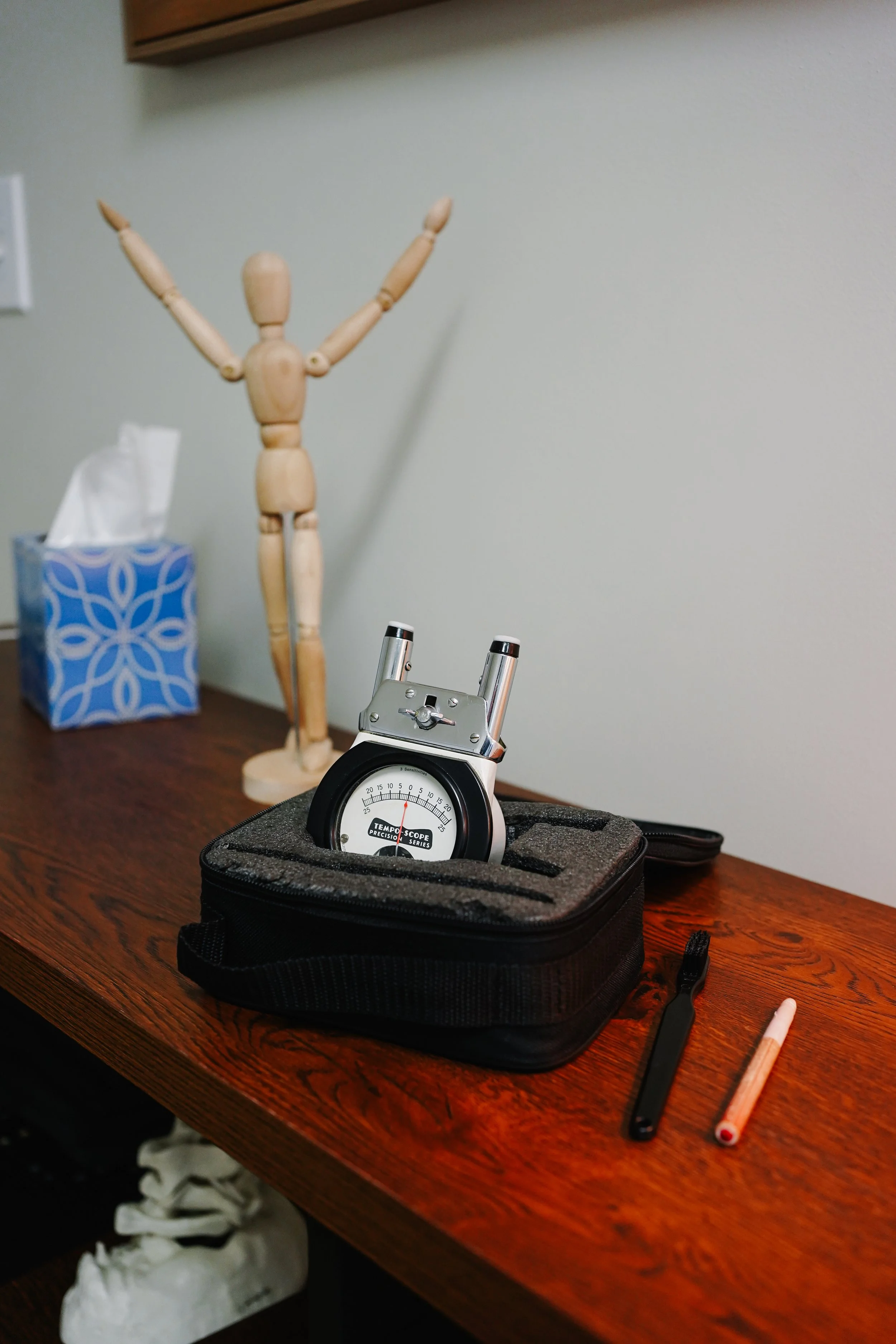 A wooden artist mannequin with arms raised stands on a desk, with a tissue box nearby. In the foreground, a black case holds a hand-held device with a dial, along with a black pen and a pink highlighter.