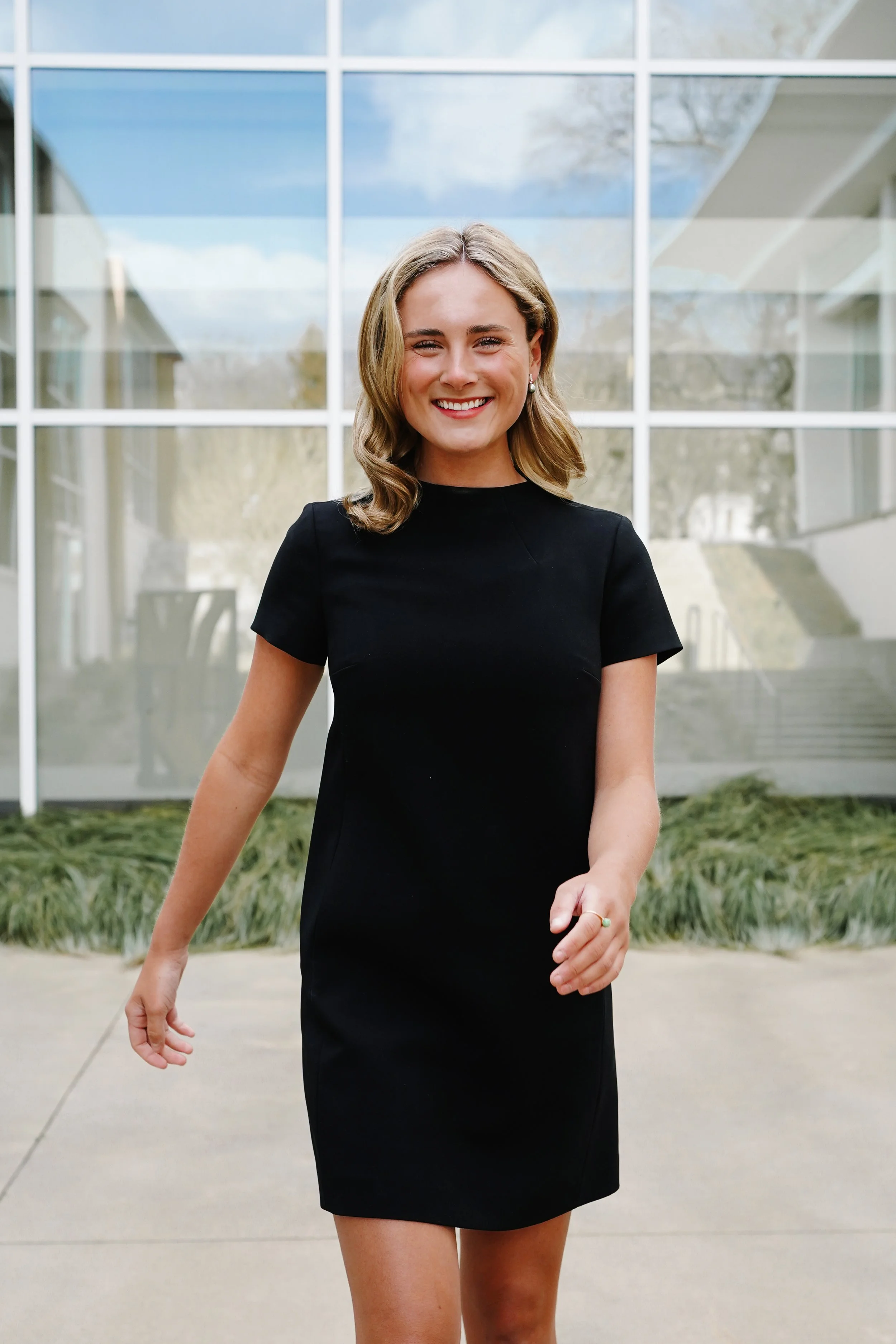 A woman with shoulder-length blonde hair smiling, walking outdoors in front of a modern glass building, wearing a black dress.