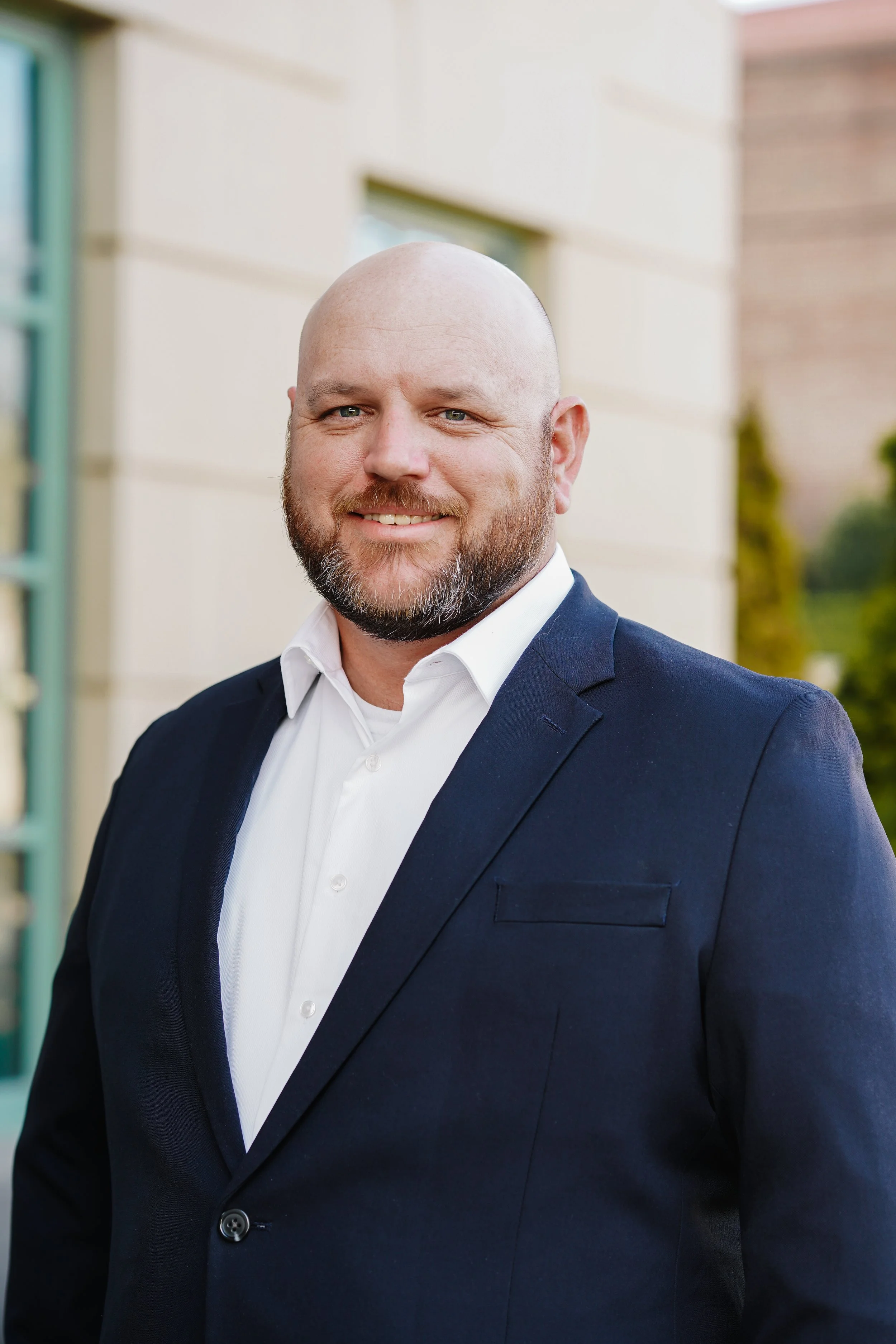 A smiling bald man with a beard in a navy blue suit and white shirt standing outdoors in front of a beige building.