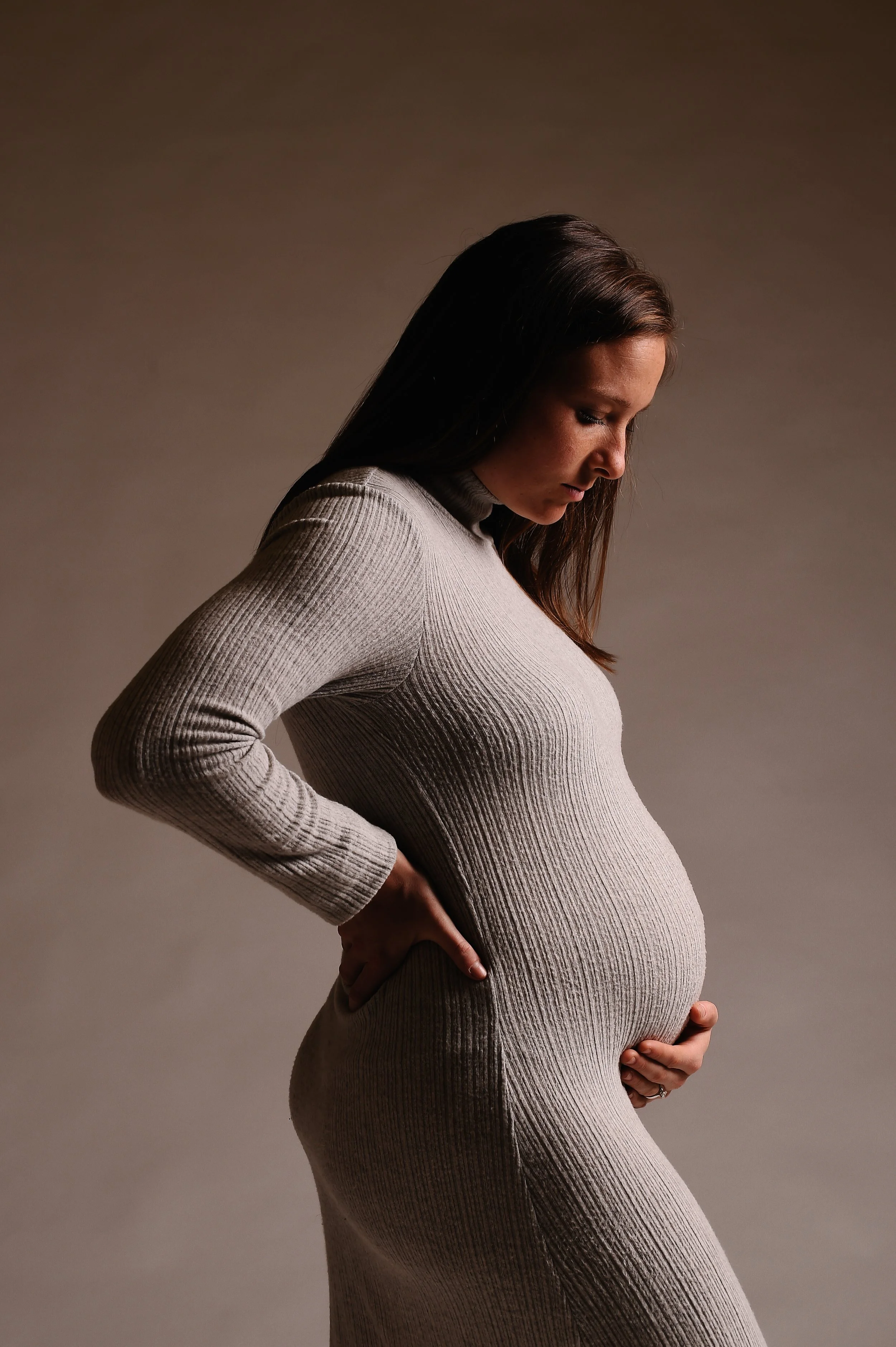 A pregnant woman in a fitted, ribbed, beige dress with long sleeves, standing in profile against a neutral background, holding her belly with one hand and resting her other hand on her back.