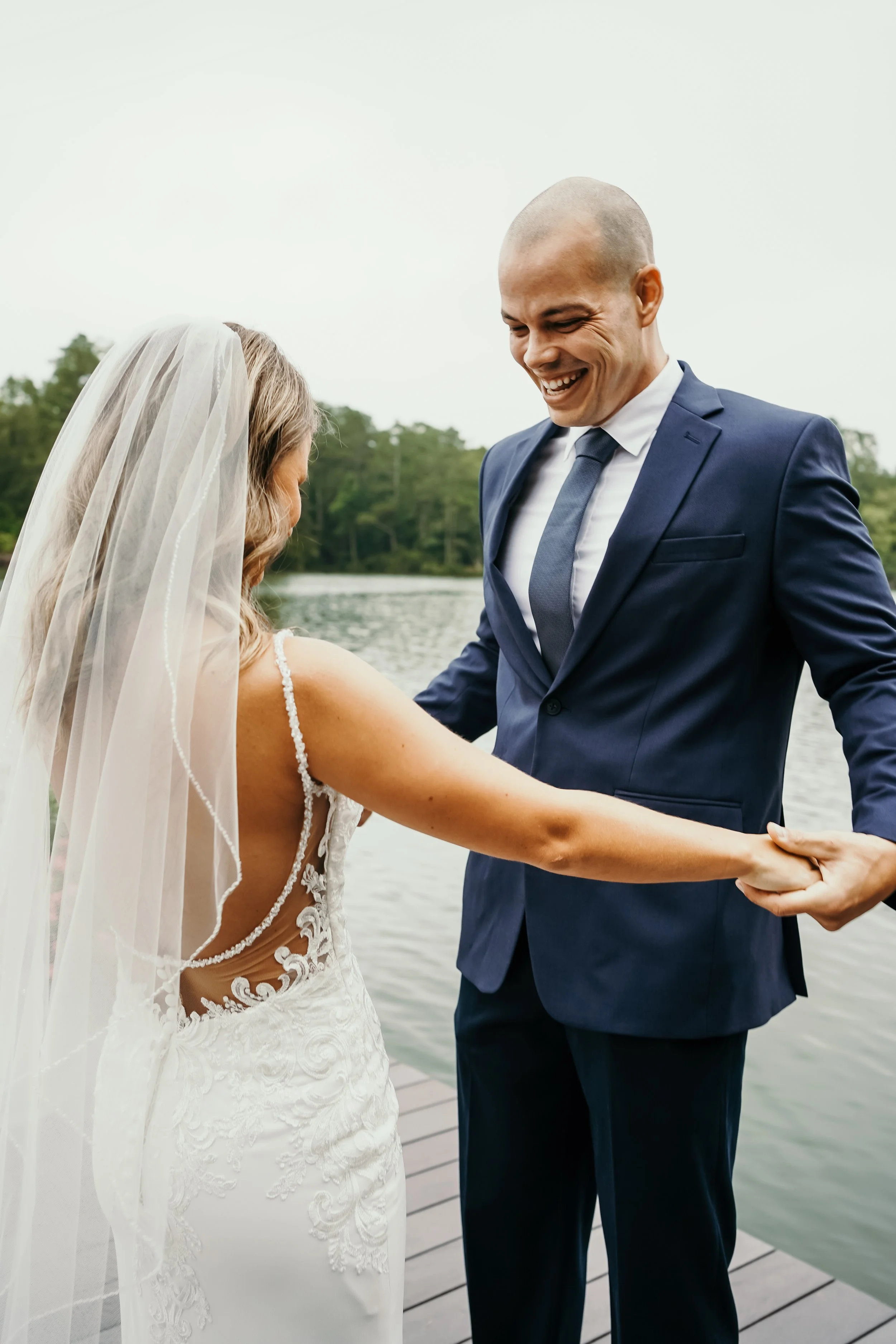 A bride and groom hold hands by a lake, smiling at each other, with the bride wearing a white wedding dress and veil and the groom in a navy suit, during their wedding ceremony.