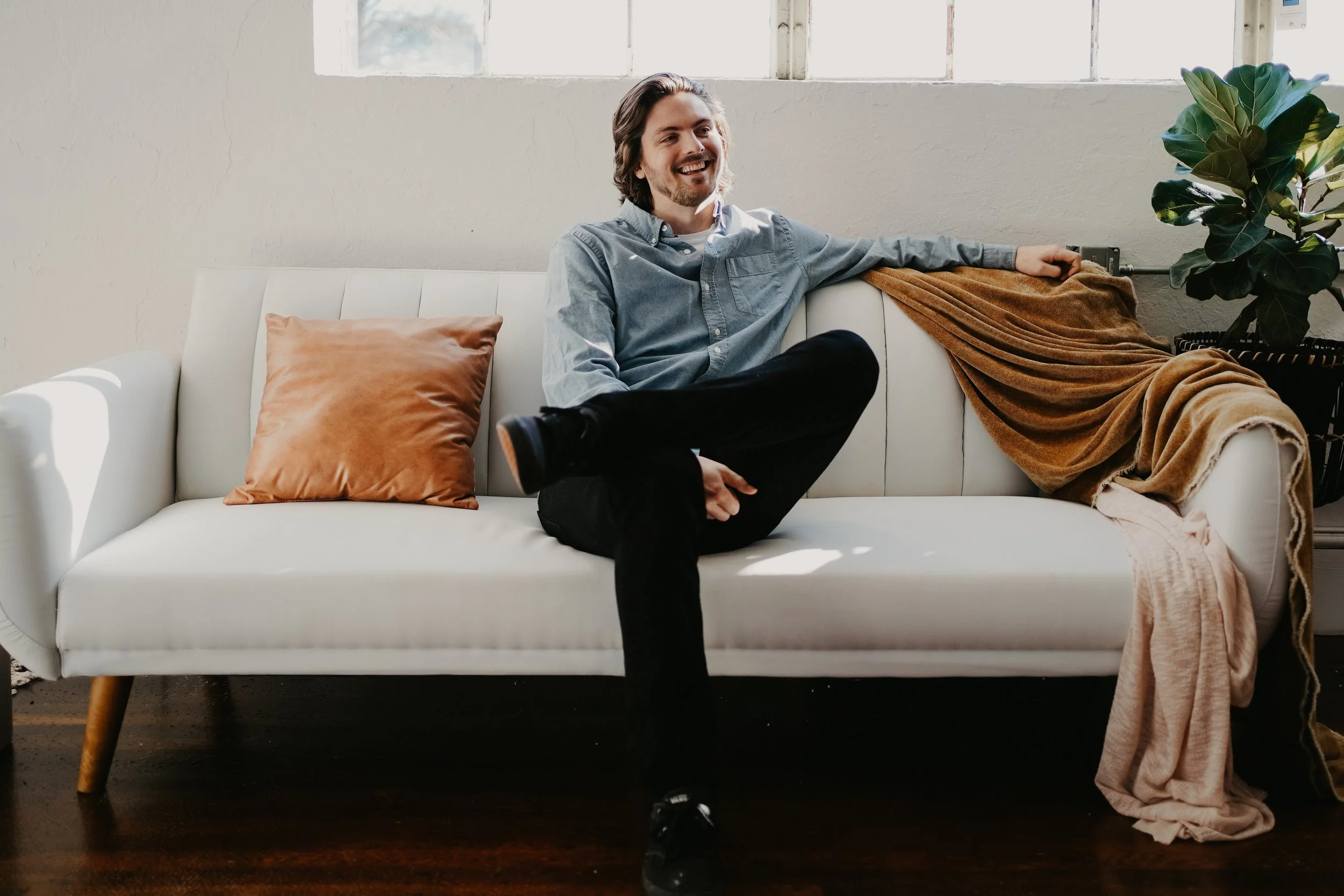 A man with shoulder-length wavy hair and a beard sitting on a white sofa, smiling happily. He is wearing a light blue button-up shirt and black pants. One leg is crossed over the other, and his right arm is resting on the back of the sofa. There is a