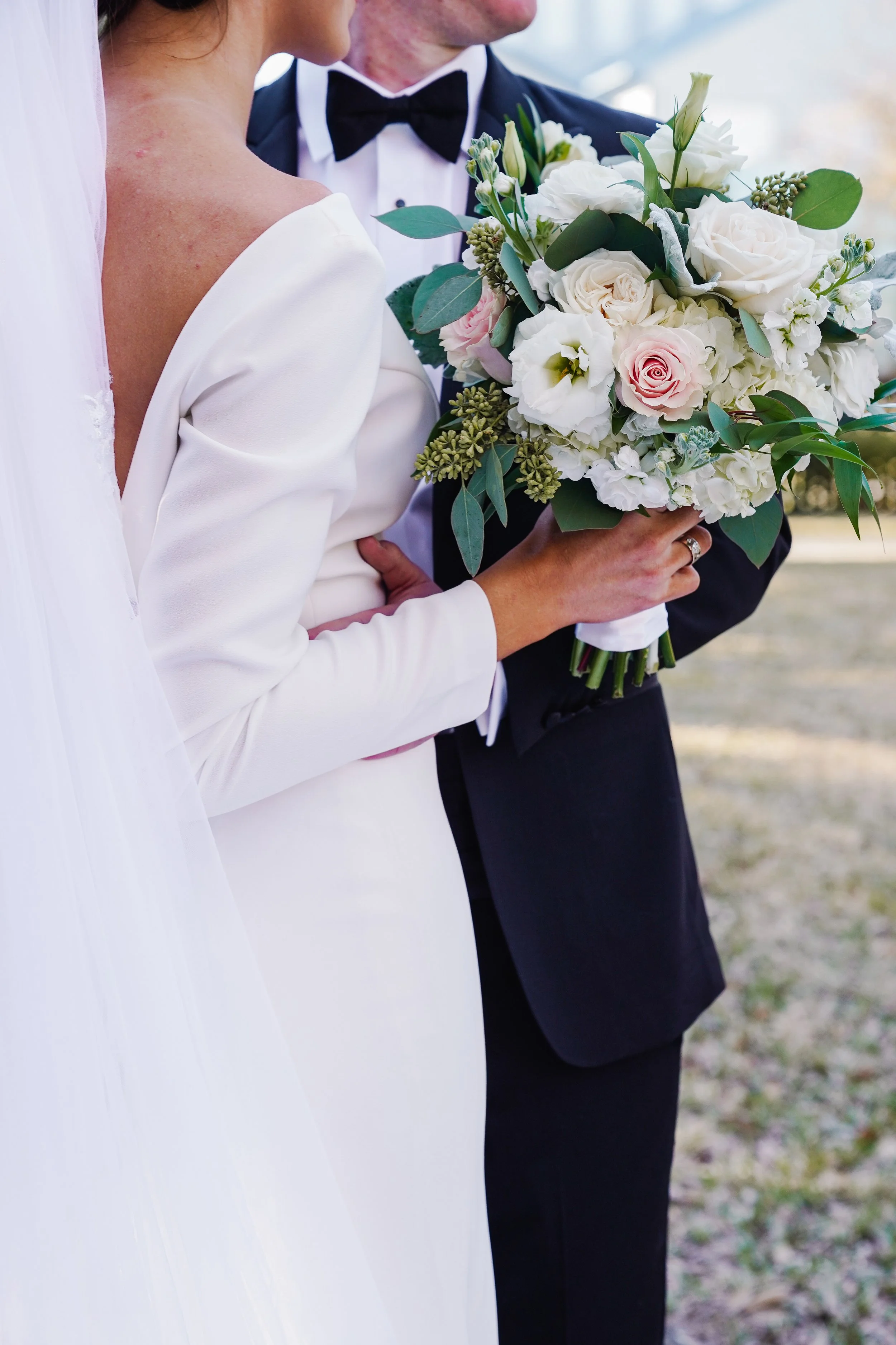 Close-up of a bride and groom holding a bouquet of white and pink roses, surrounded by greenery, during their wedding ceremony.