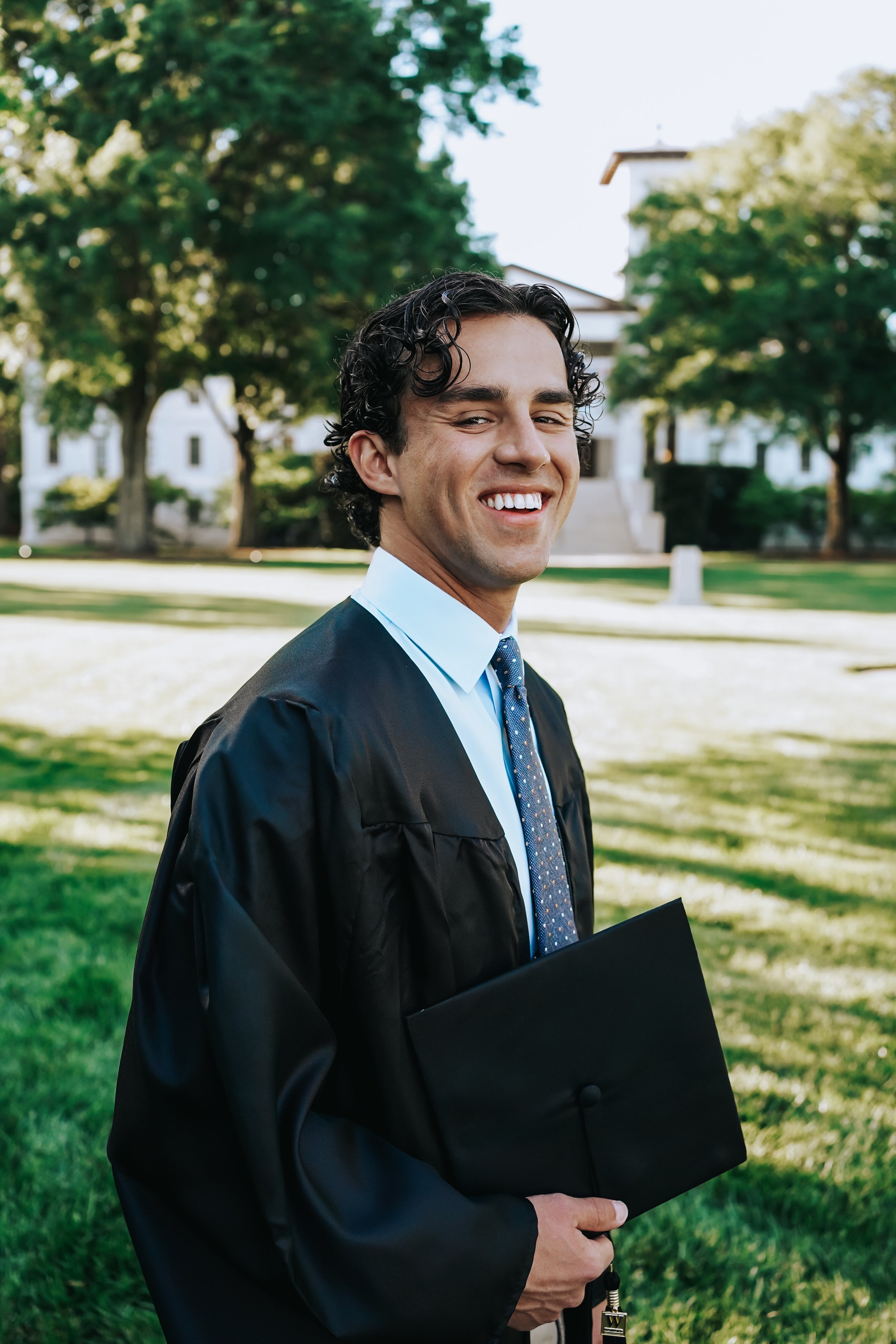 A young man in a graduation gown holding a diploma, smiling outdoors on a sunny day with trees and a building in the background.